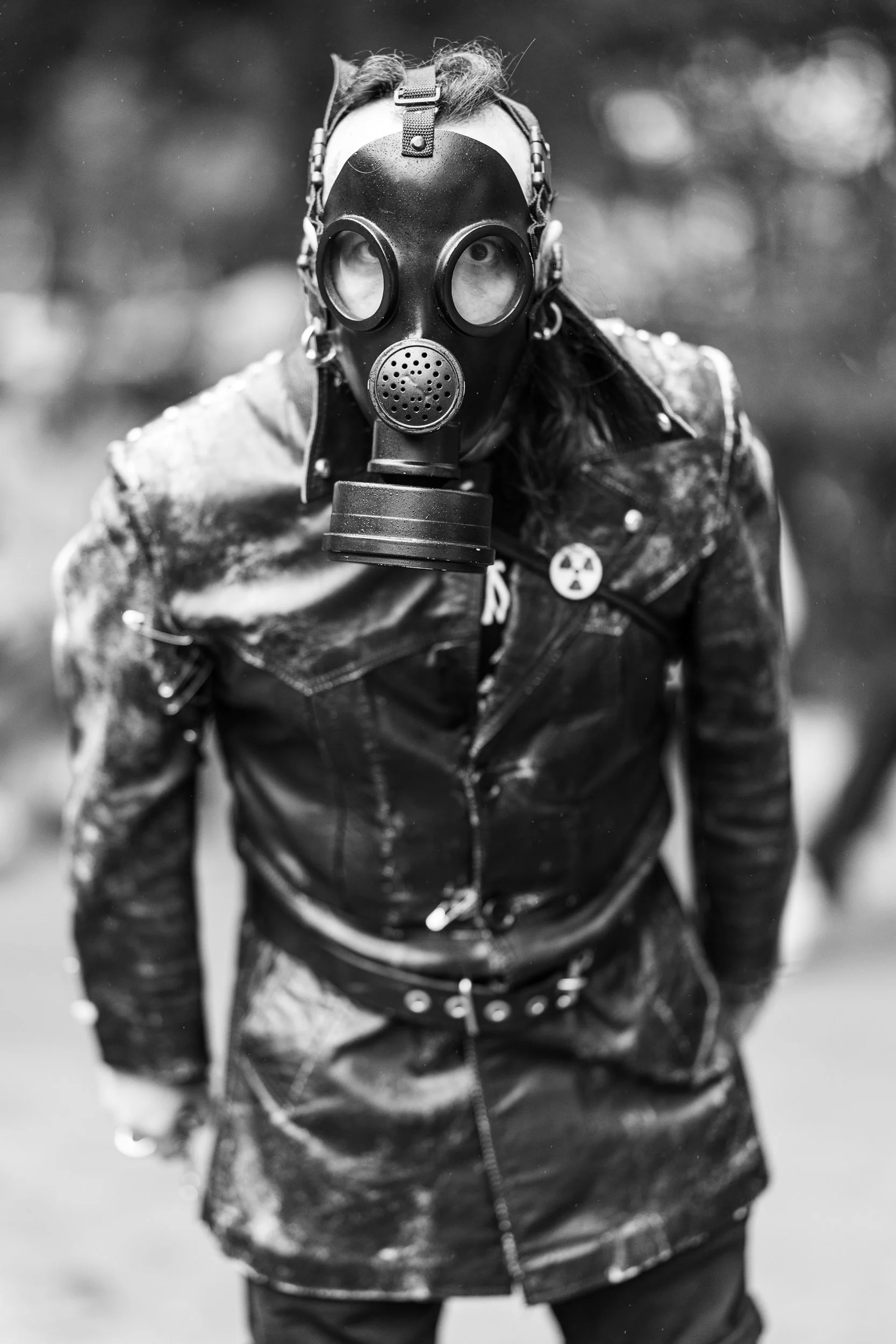 A person wearing a gas mask and a leather jacket with a radioactive symbol patch, standing outdoors in a blurred natural environment.