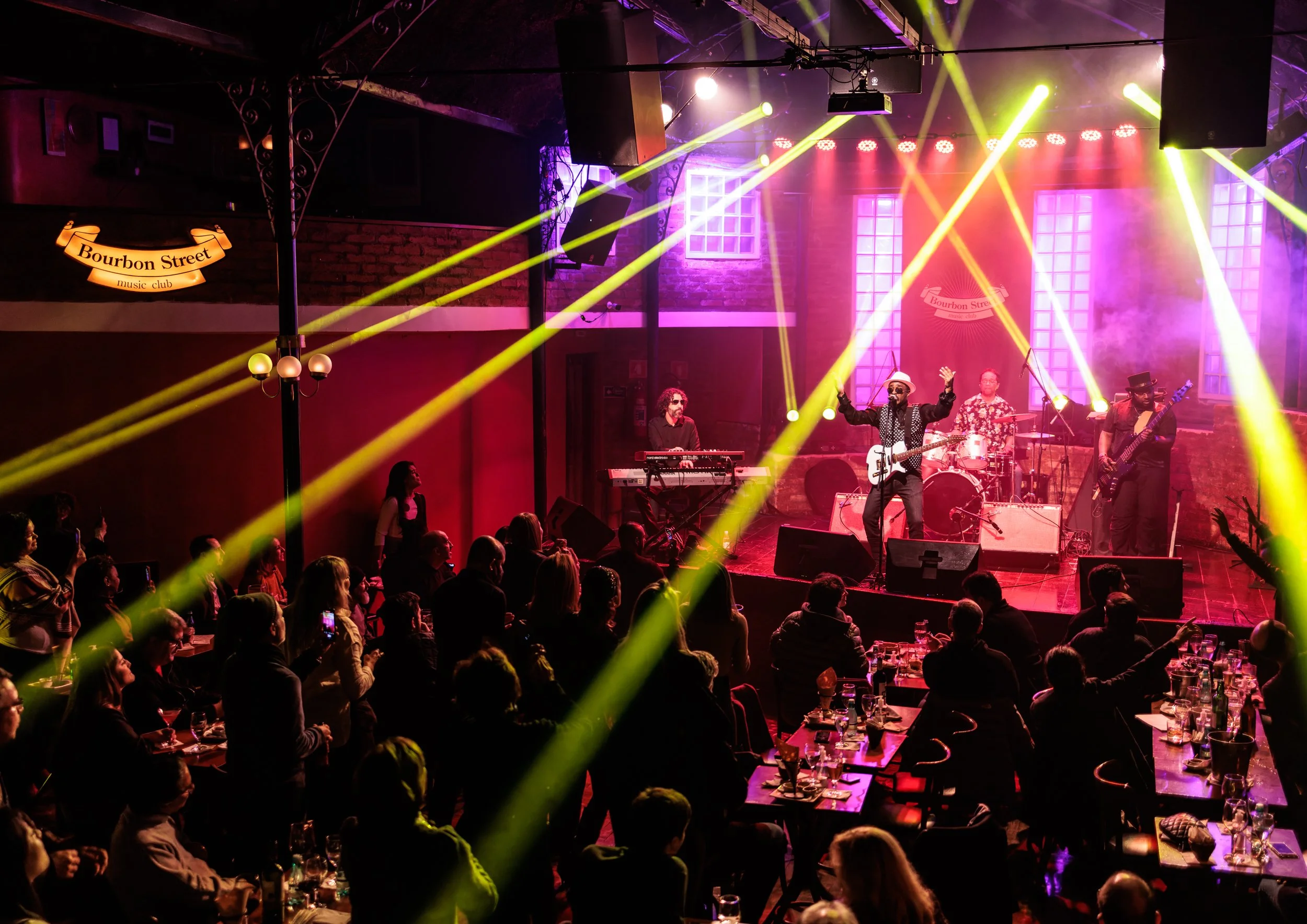 A live music performance at Bourbon Street music club with a band playing on stage, colorful stage lights, and an audience seated at tables.