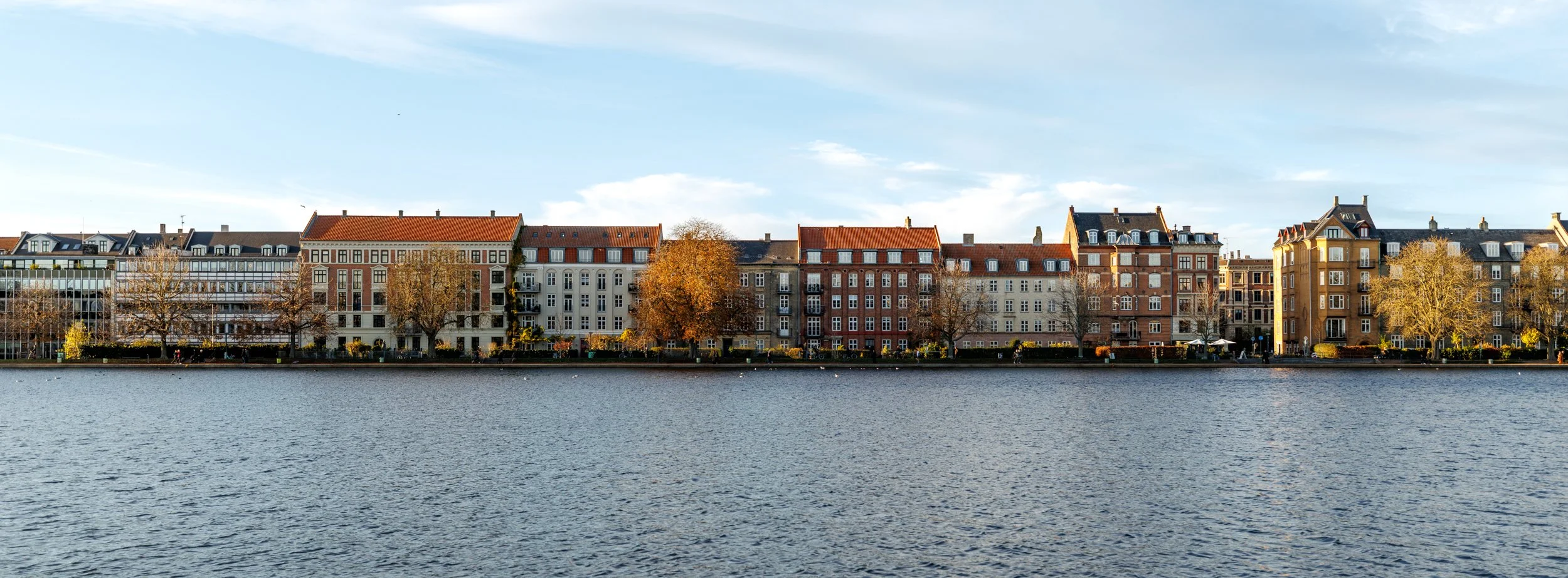A row of colorful European-style buildings alongside a river, with trees in front and a partly cloudy sky above.