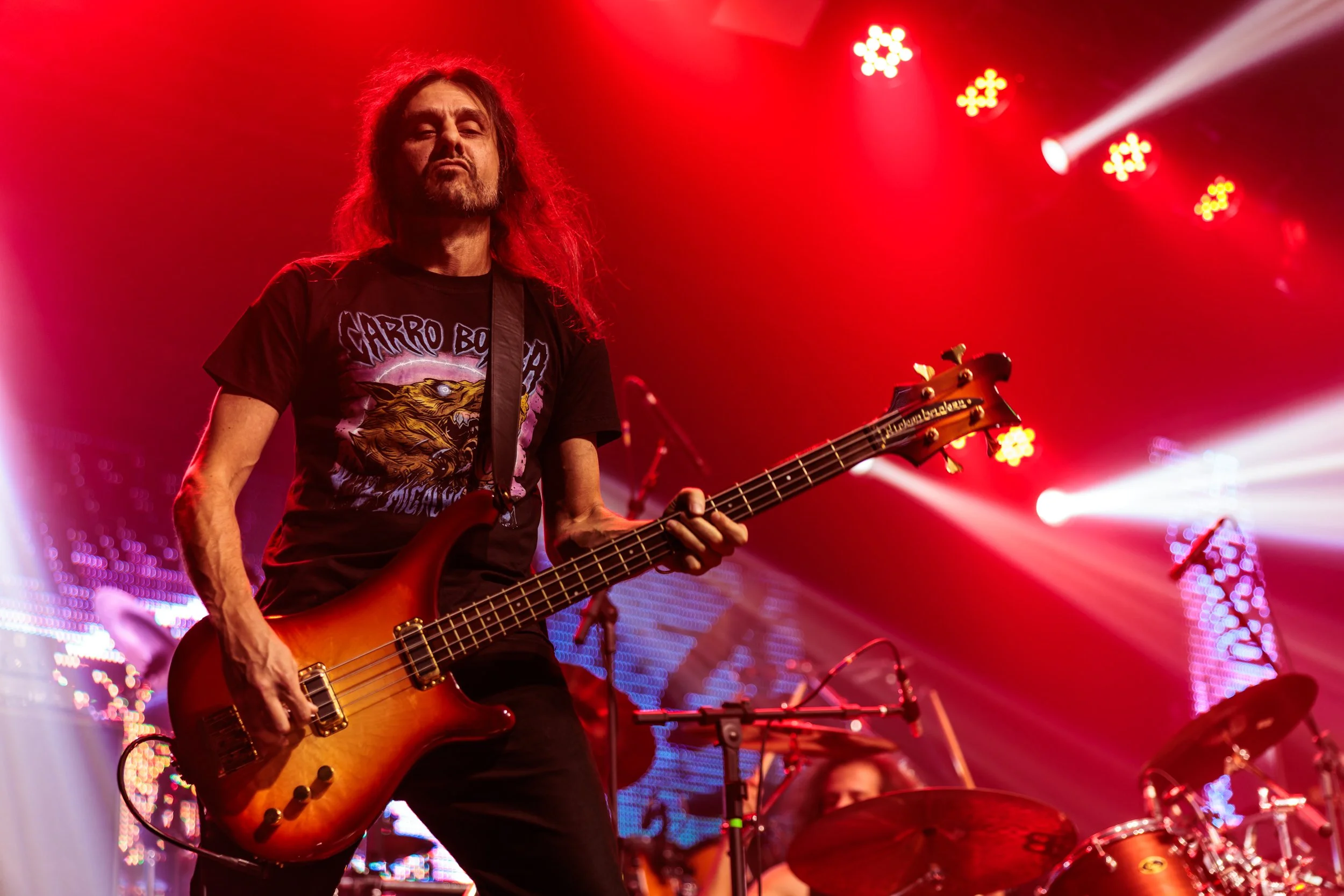 A musician with long hair and a beard playing an electric bass guitar on stage during a concert. The background is illuminated with red stage lighting and LED screens.