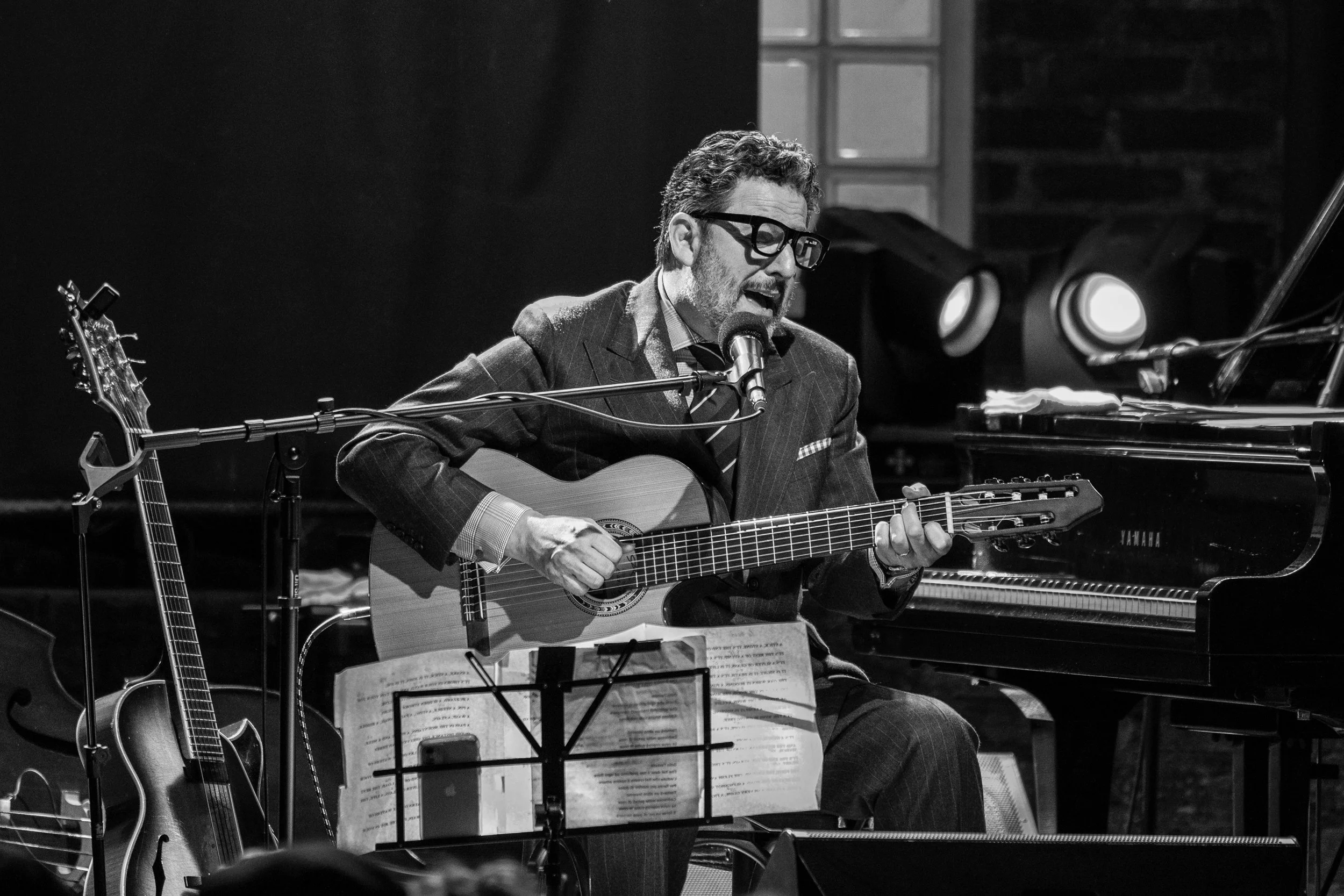 A man with glasses and a beard playing an acoustic guitar and singing into a microphone during a performance in a dimly lit room.