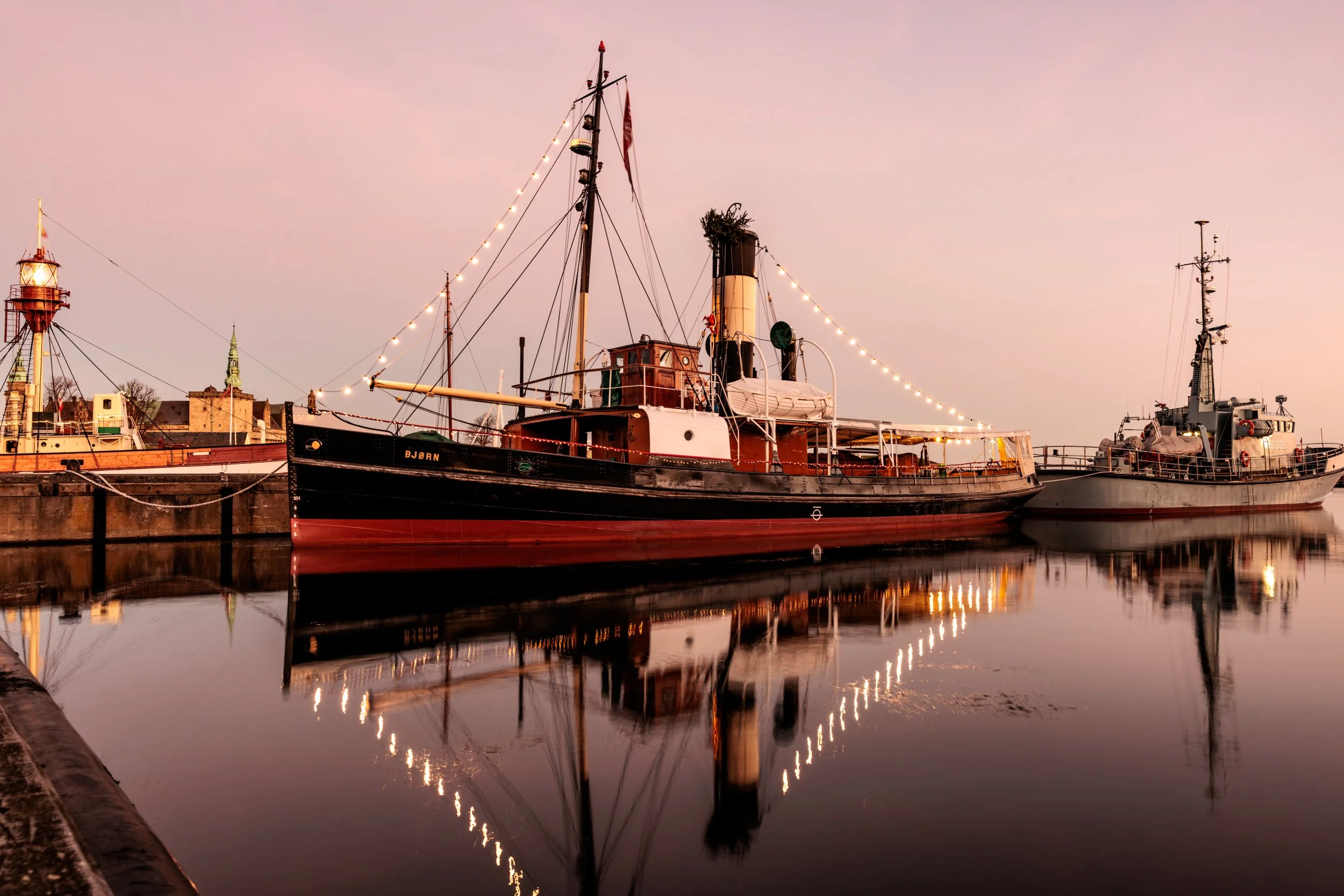 A calm harbor scene at sunset with two boats docked, string lights hanging along the boats, and historic buildings in the background, reflected on the water.