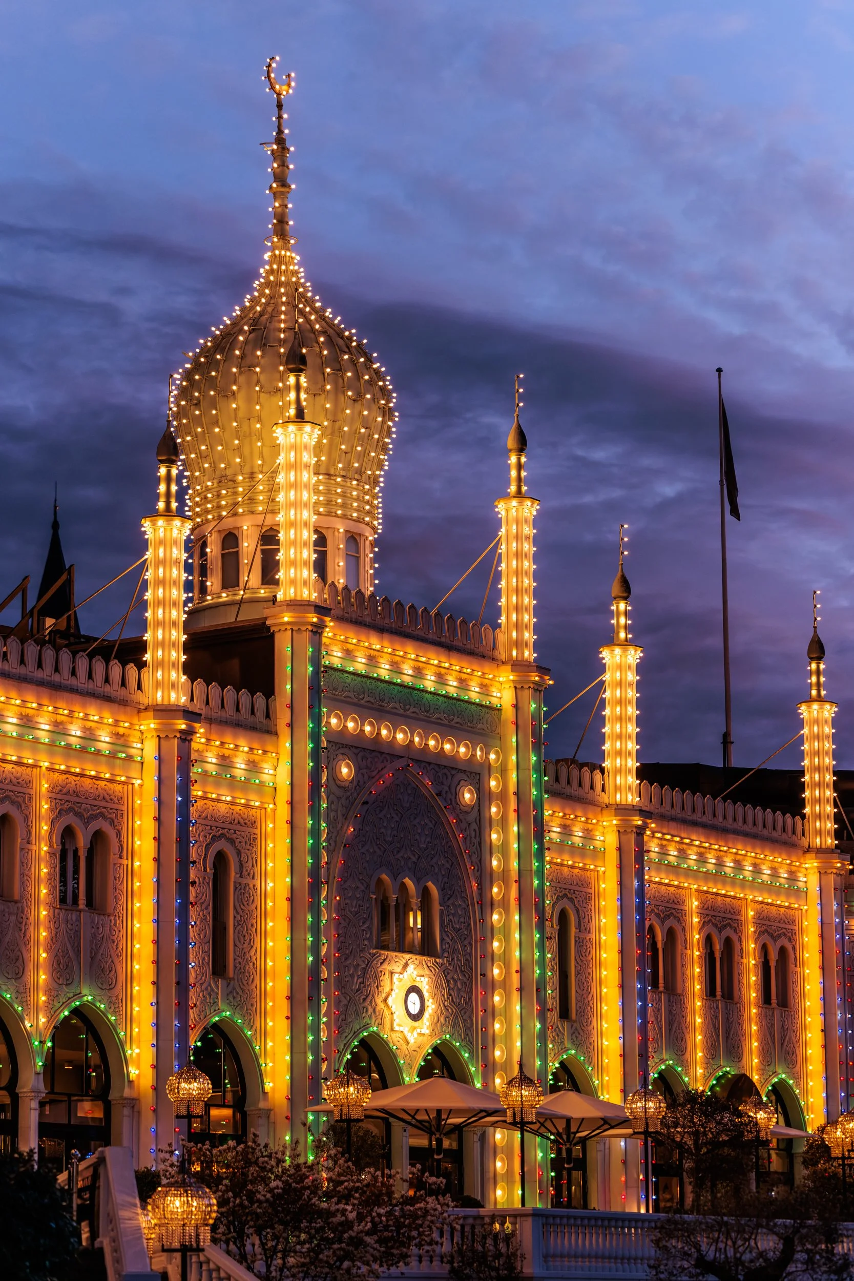 A beautifully lit castle-like building decorated with string lights in warm yellow, green, red, and blue, with a large clock on the front, set against a twilight sky with dark clouds.