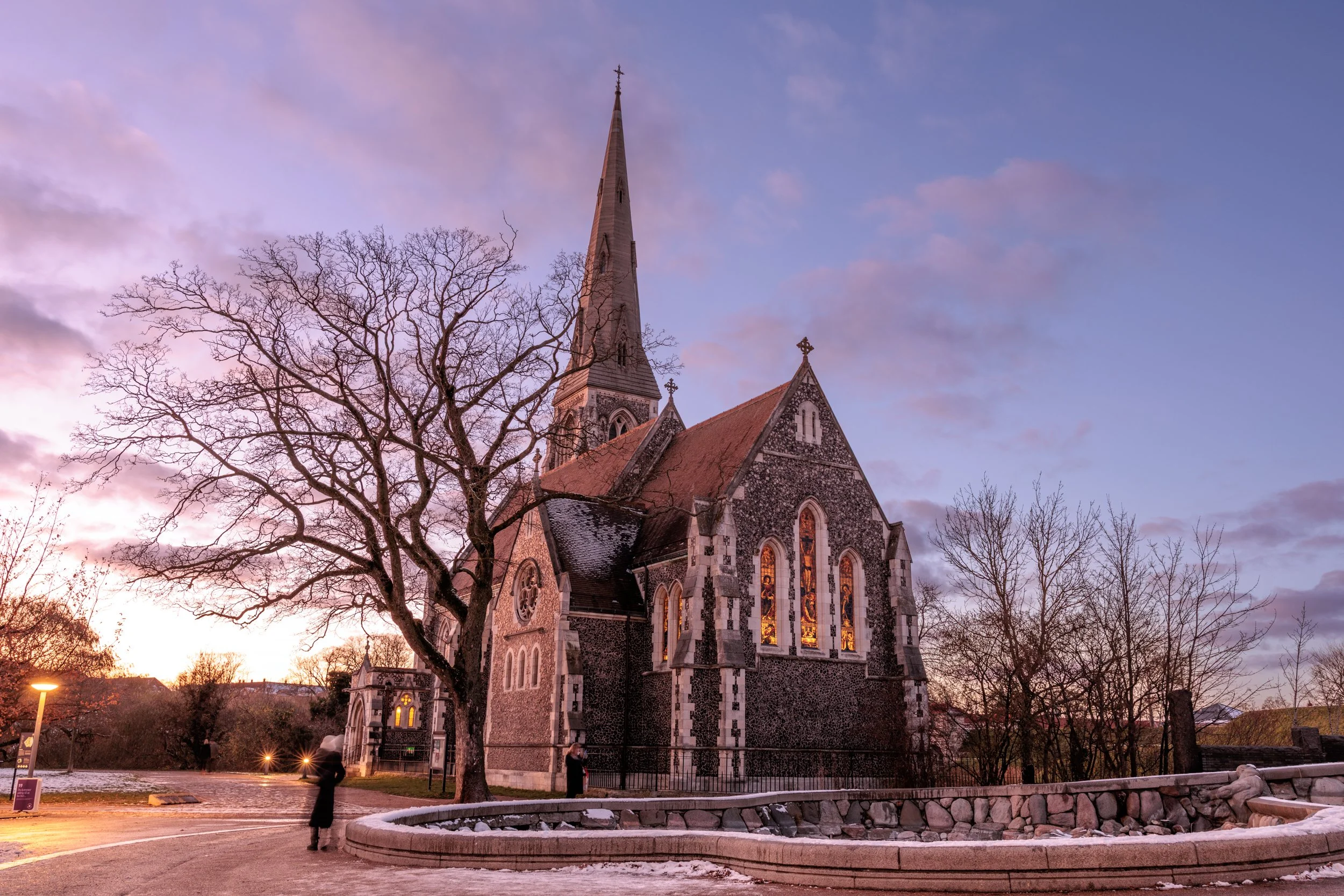 A stone church with a tall steeple, stained glass windows illuminated from inside, and leafless trees in front, set against a purple and blue sky at dusk.