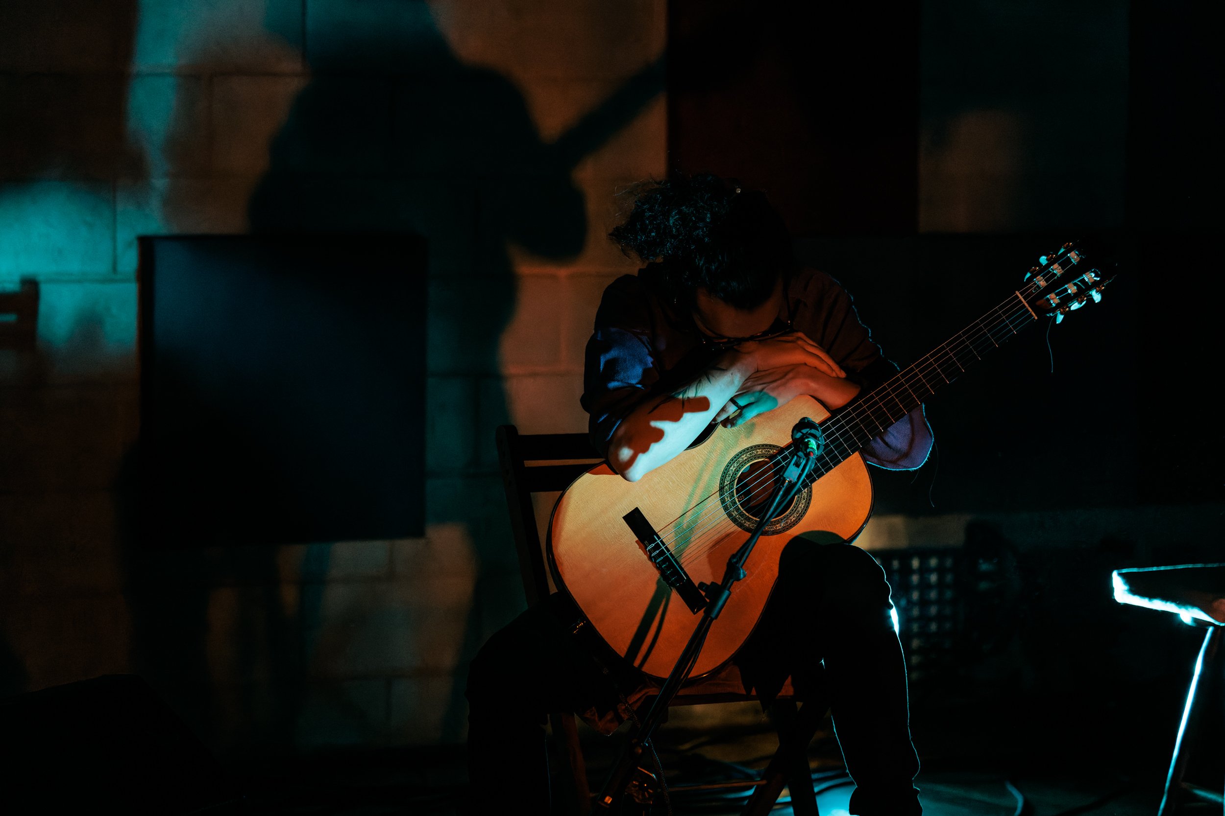 A person with curly hair playing an acoustic guitar on a dimly lit stage, with a microphone positioned in front of them, and a shadow cast on the wall behind.