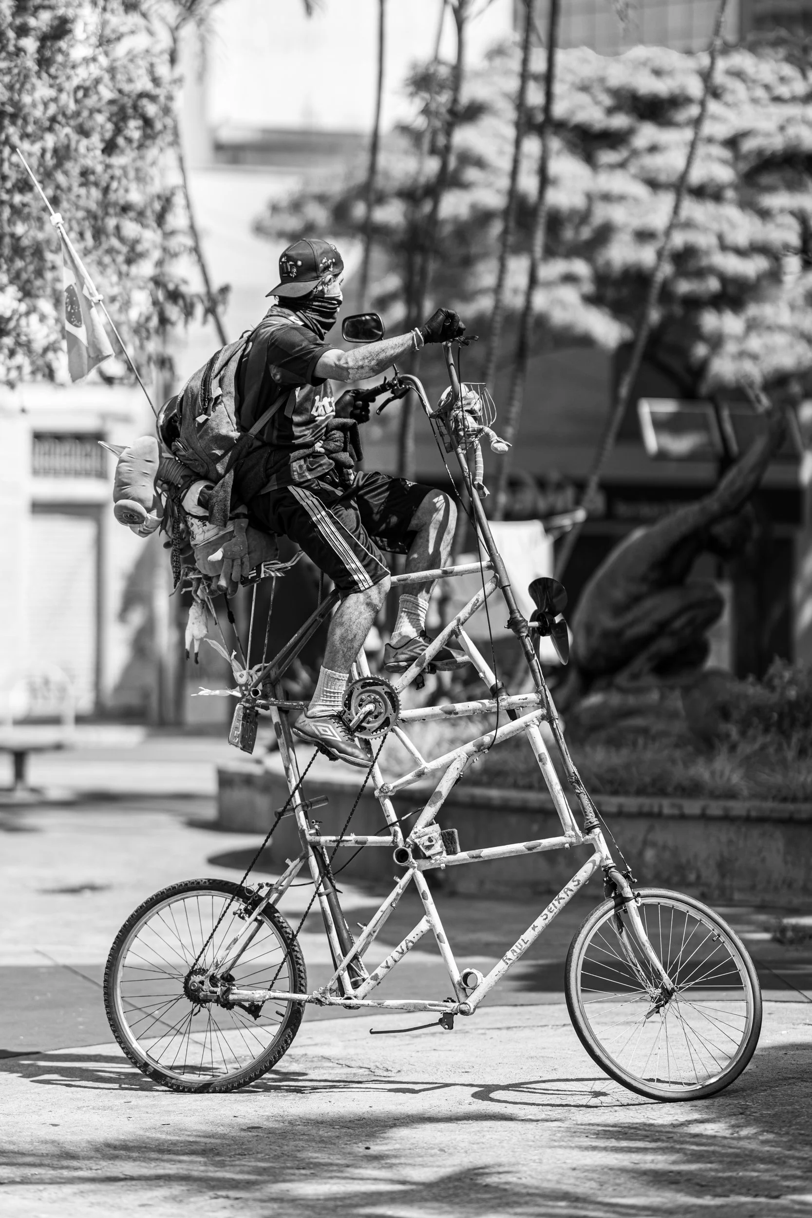 A person wearing a hat, face cover, and casual clothes rides a tall, custom-built bicycle with an extra frame, possibly a tandem or artistic bike, in an outdoor urban setting with trees and buildings in the background.