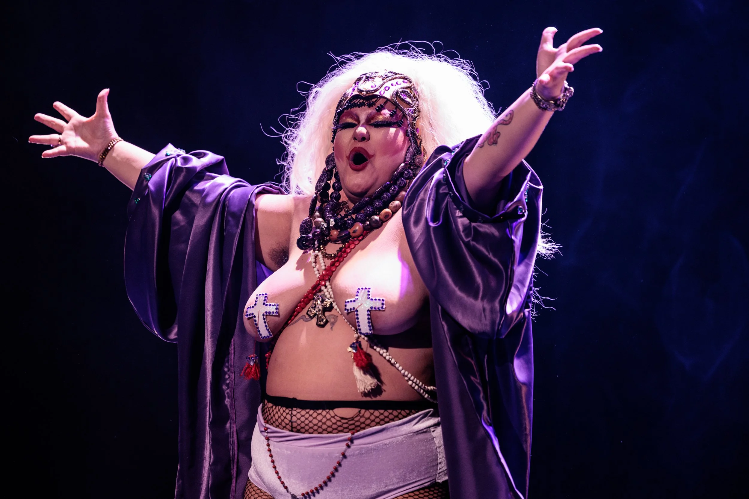 Performer with curly blonde hair wearing a purple satin jacket, fishnet stockings, and layered jewelry, with arms raised and eyes closed, on stage with dark background.