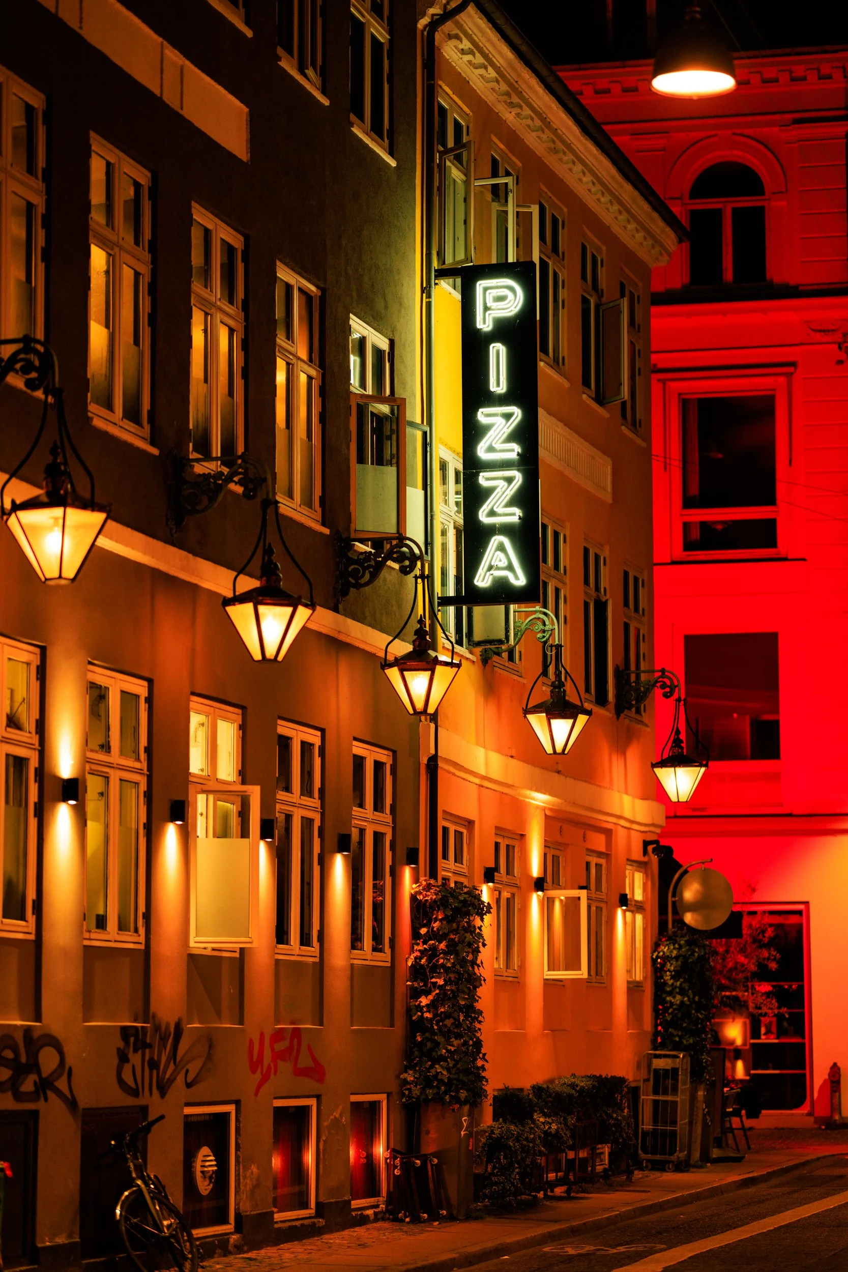 Night scene of a city street with illuminated lamps and a vertical neon sign that reads 'PIZZA'. The building facades are lit with warm yellow and red lighting, and windows are open with some showing interior light. Graffiti is visible on the lower p