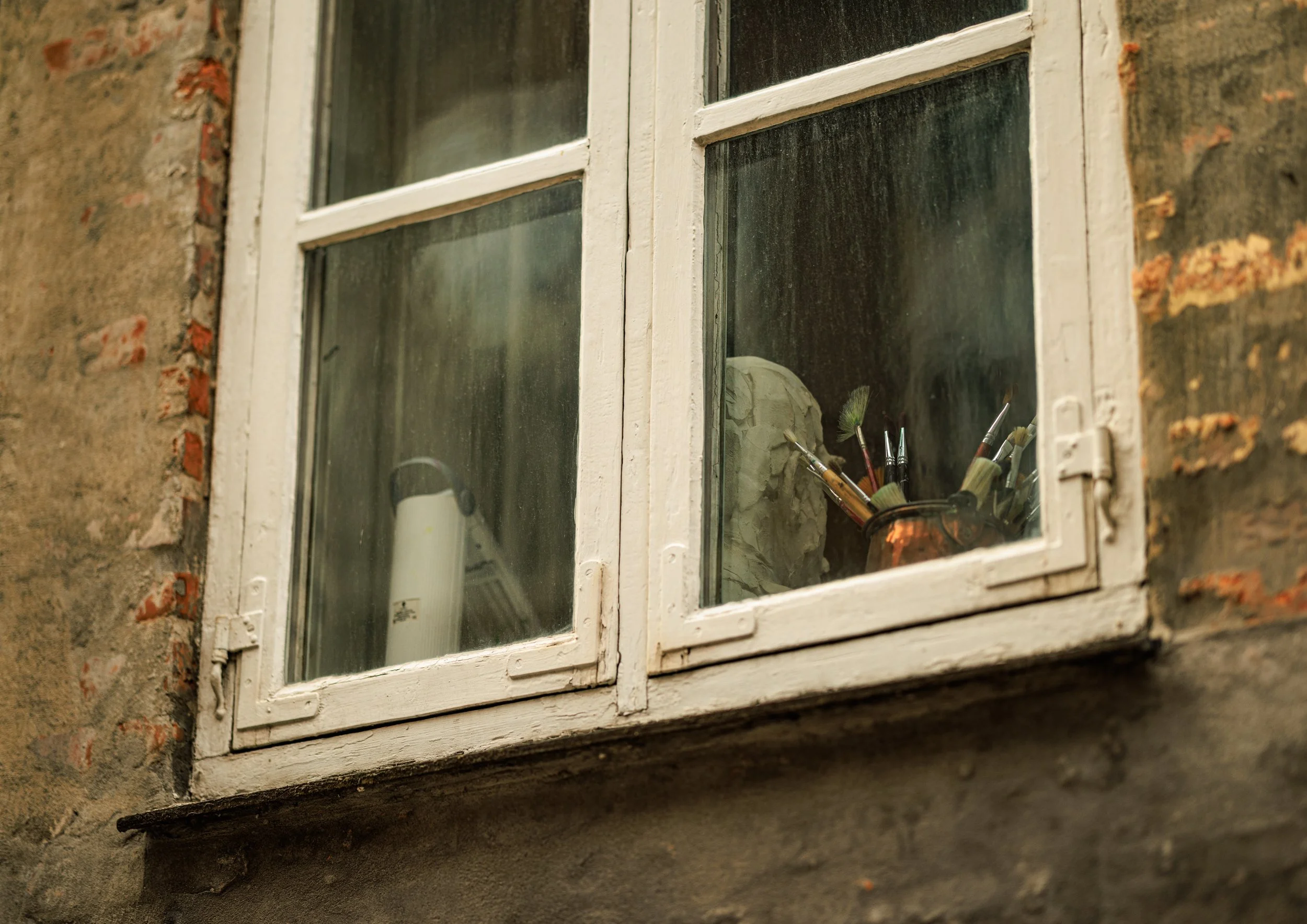 A close-up of an old brick wall with a white wooden-framed window, through which a display of paintbrushes and cleaning supplies on a shelf inside a building are visible.