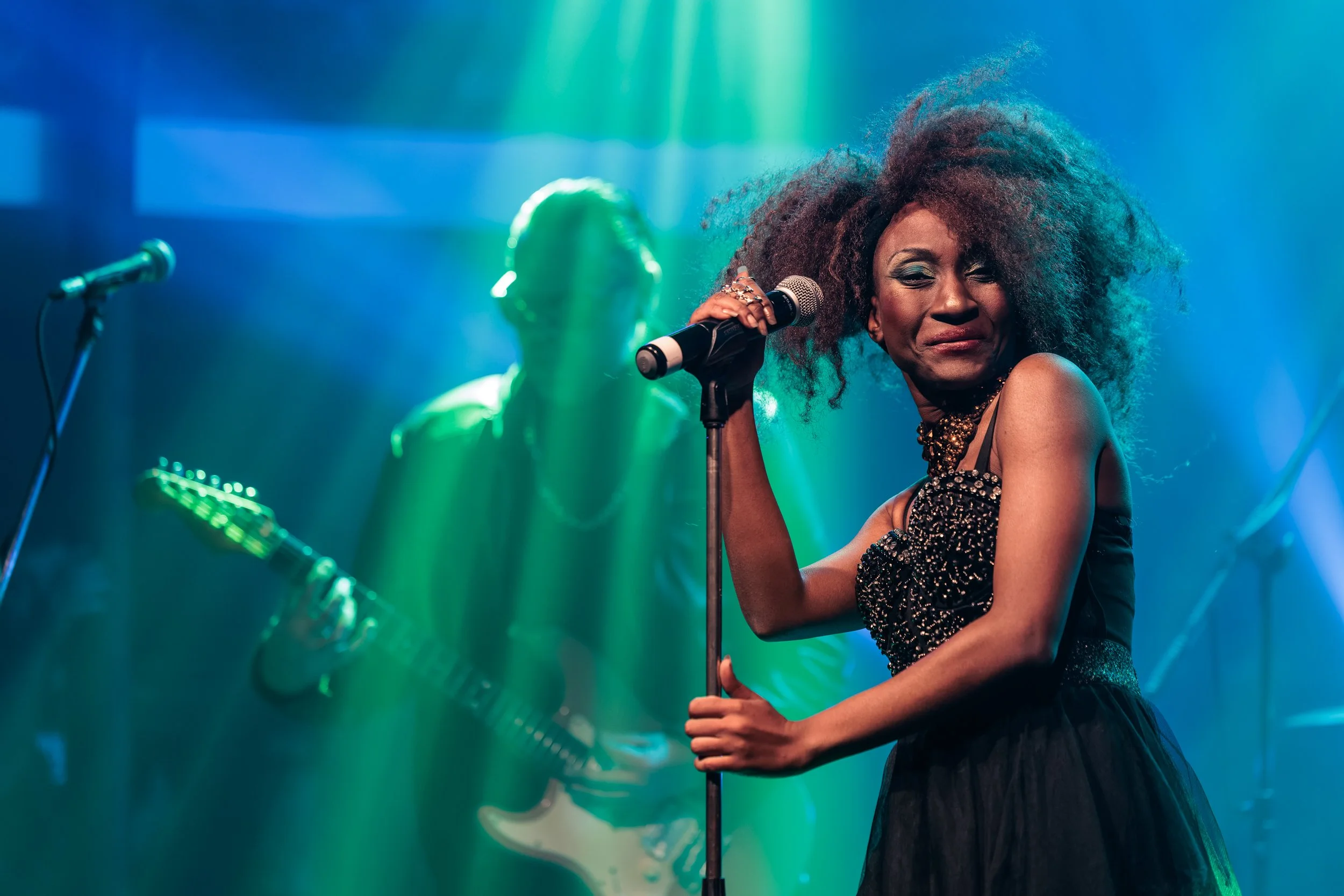 A female singer with curly hair in a black dress, holding a microphone on stage with colorful lighting, and a guitarist in the background.