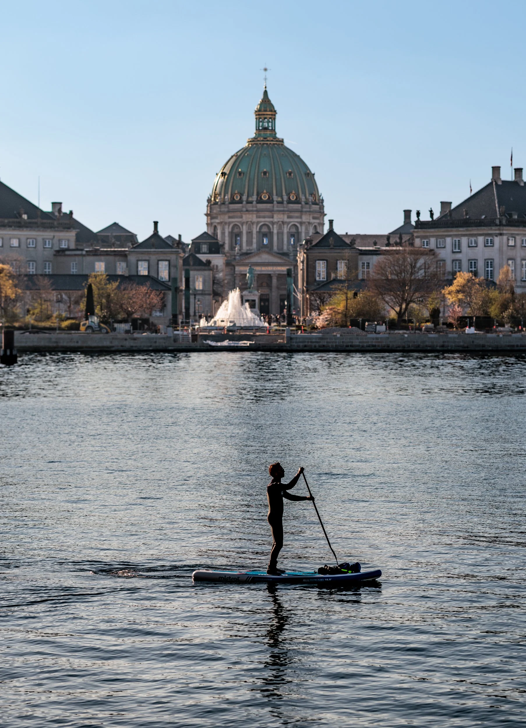 A person paddling on a stand-up paddleboard in a river with a fountain, historic buildings, and a domed government building in the background during the day.