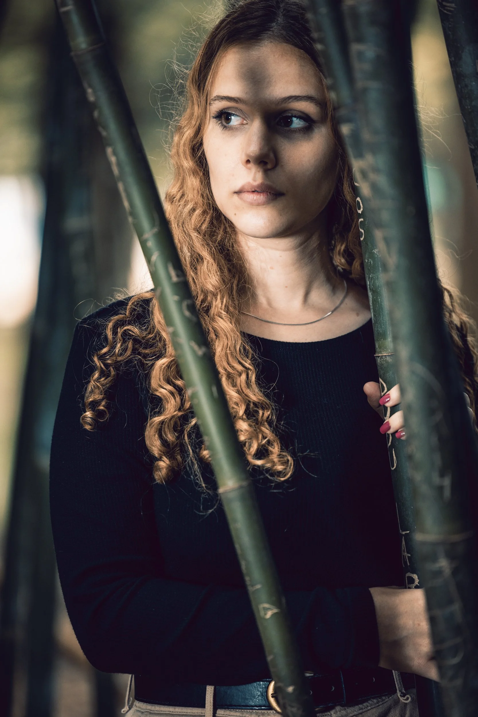 A young woman with long, curly hair and light skin standing behind black metal bars, looking to the side with a neutral expression, outdoors in natural light.