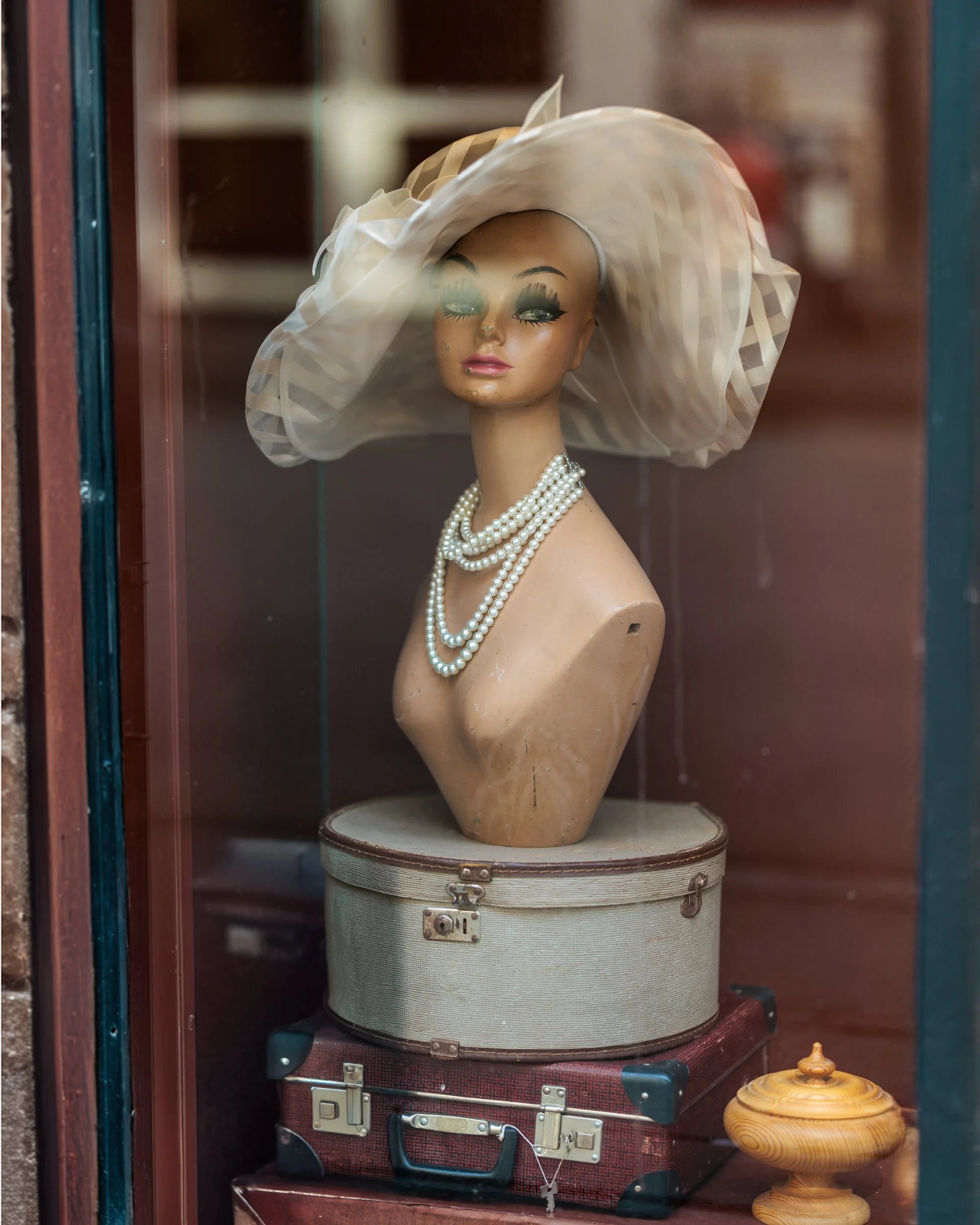 A mannequin bust wearing a large white wide-brimmed hat and pearl necklace, displayed inside a glass cabinet, with a brown suitcase and a wooden decorative object nearby.