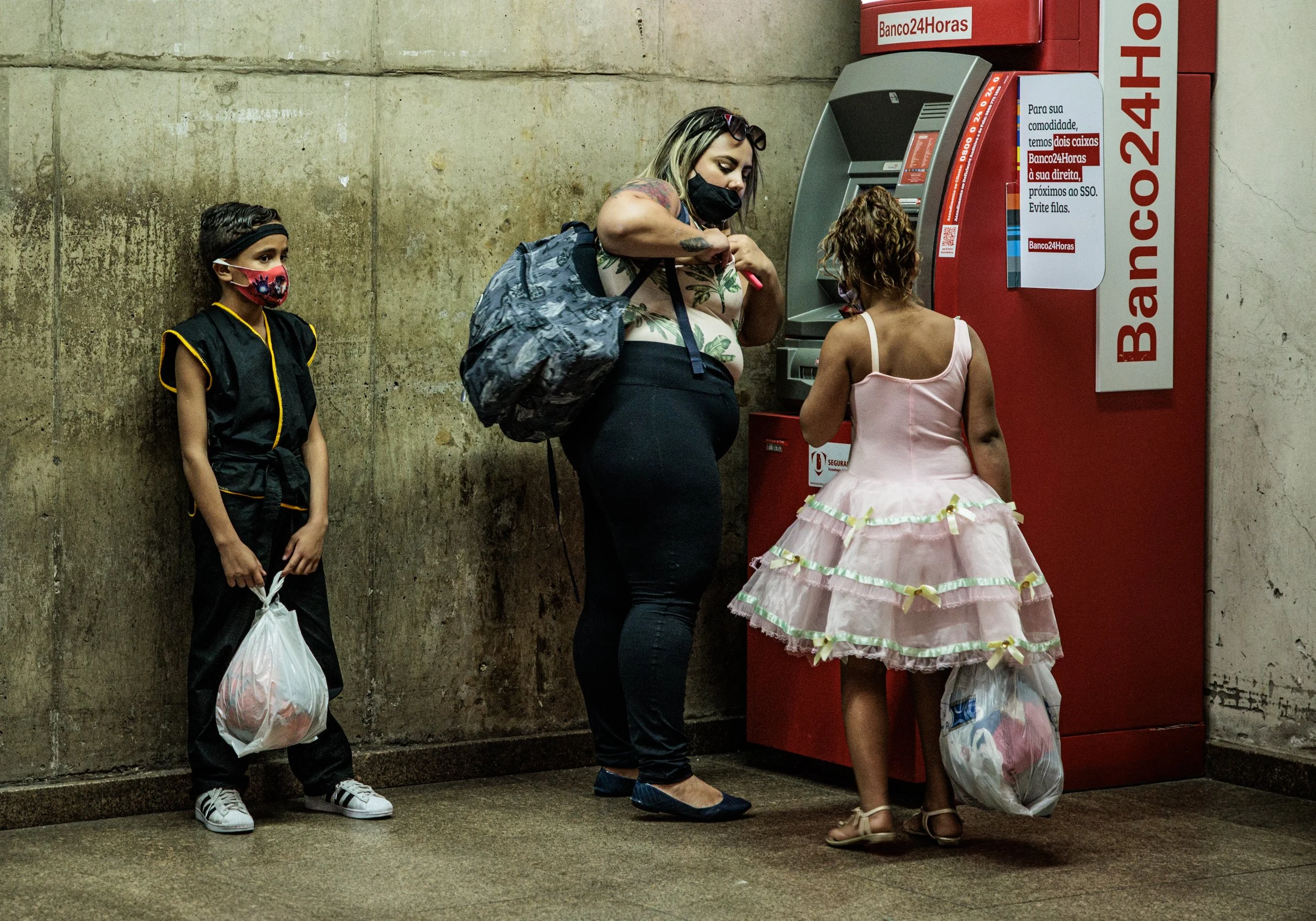 A woman and two young girls standing near an ATM machine in a public space. The woman is looking at the ATM, wearing a black face mask, a sleeveless top with a floral pattern, and carrying a backpack. One girl is dressed in a dark martial arts unifor