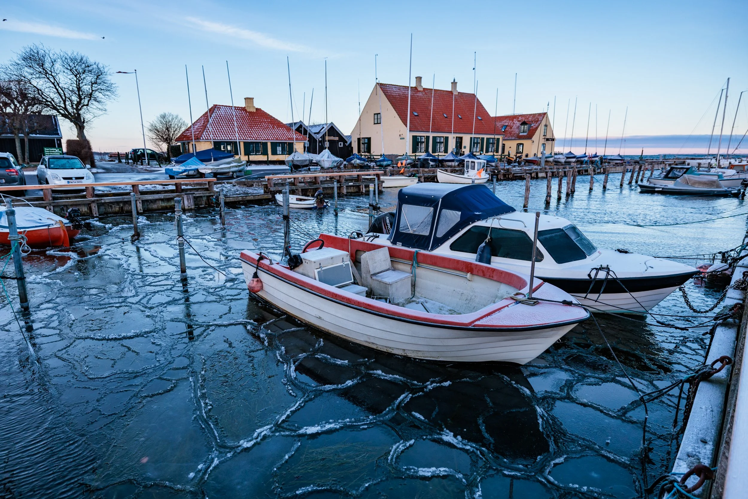 A marina with boats docked along the pier in winter, snow on the ground and rooftops, and a few leafless trees, under a clear sky.