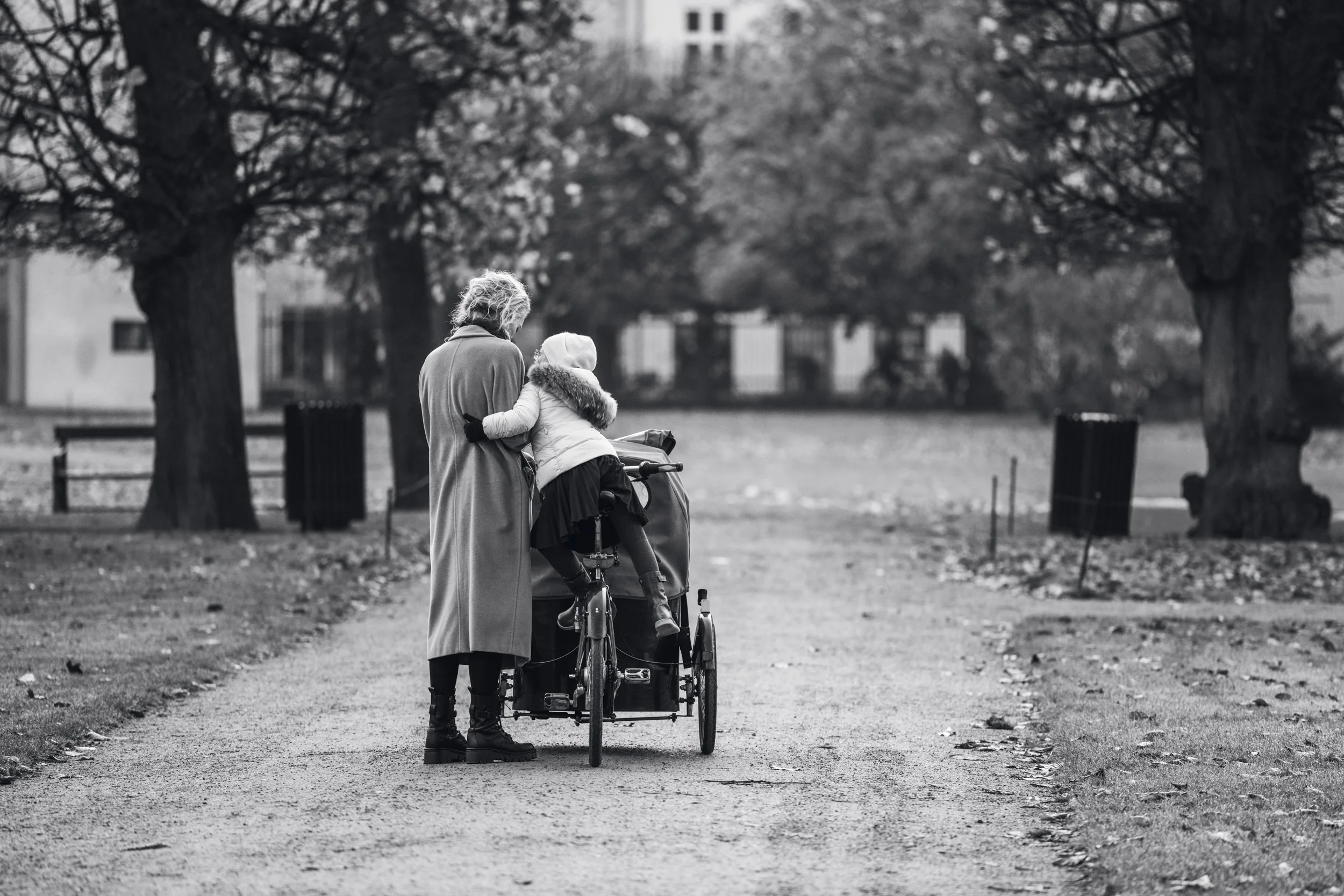 An elderly woman standing with her back to the camera, wearing a long coat and boots, assisting a young girl with her bicycle in a park surrounded by trees.
