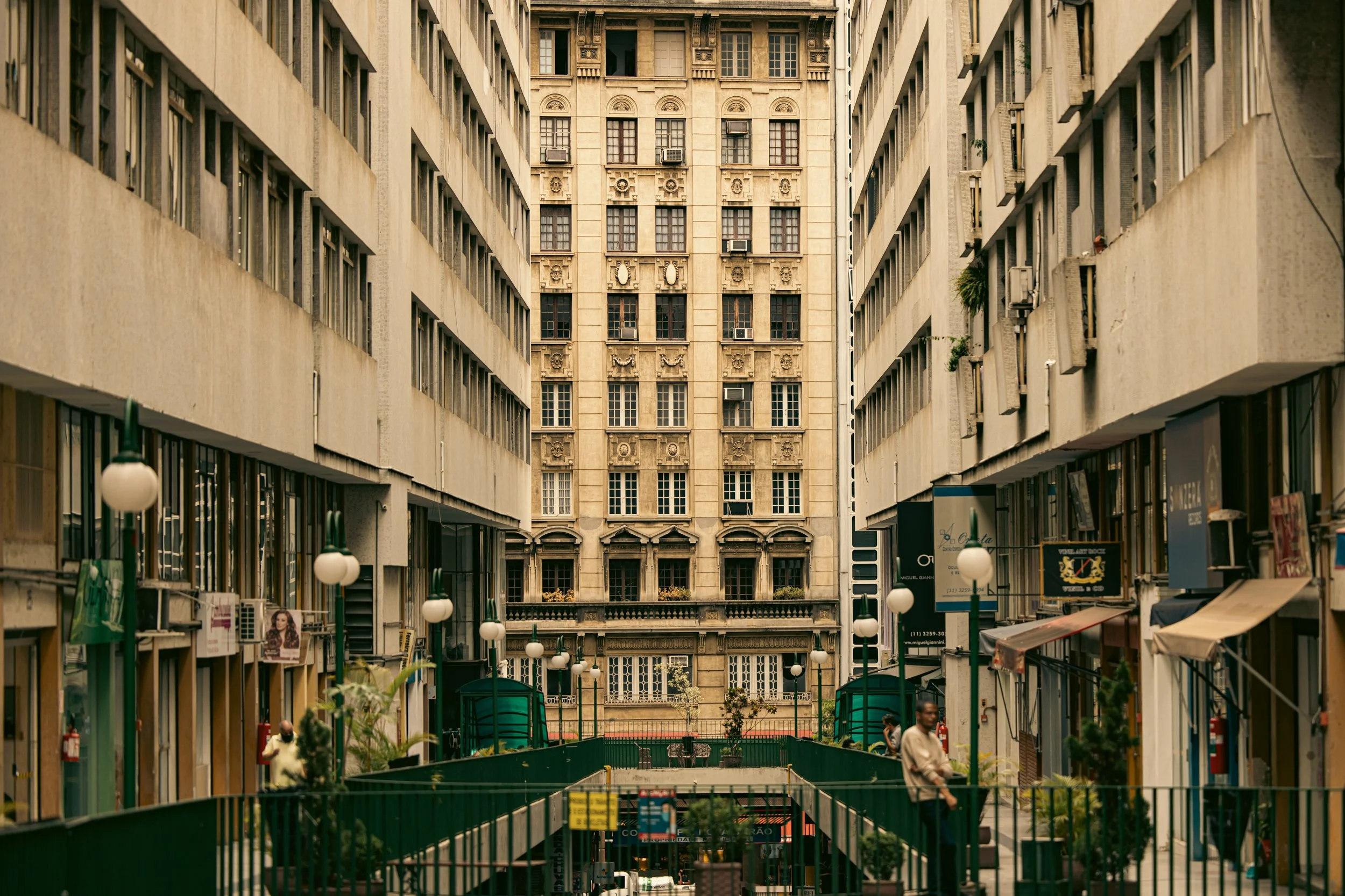 City street with tall buildings on both sides, storefronts at street level, and decorative street lamps hanging over the sidewalk.