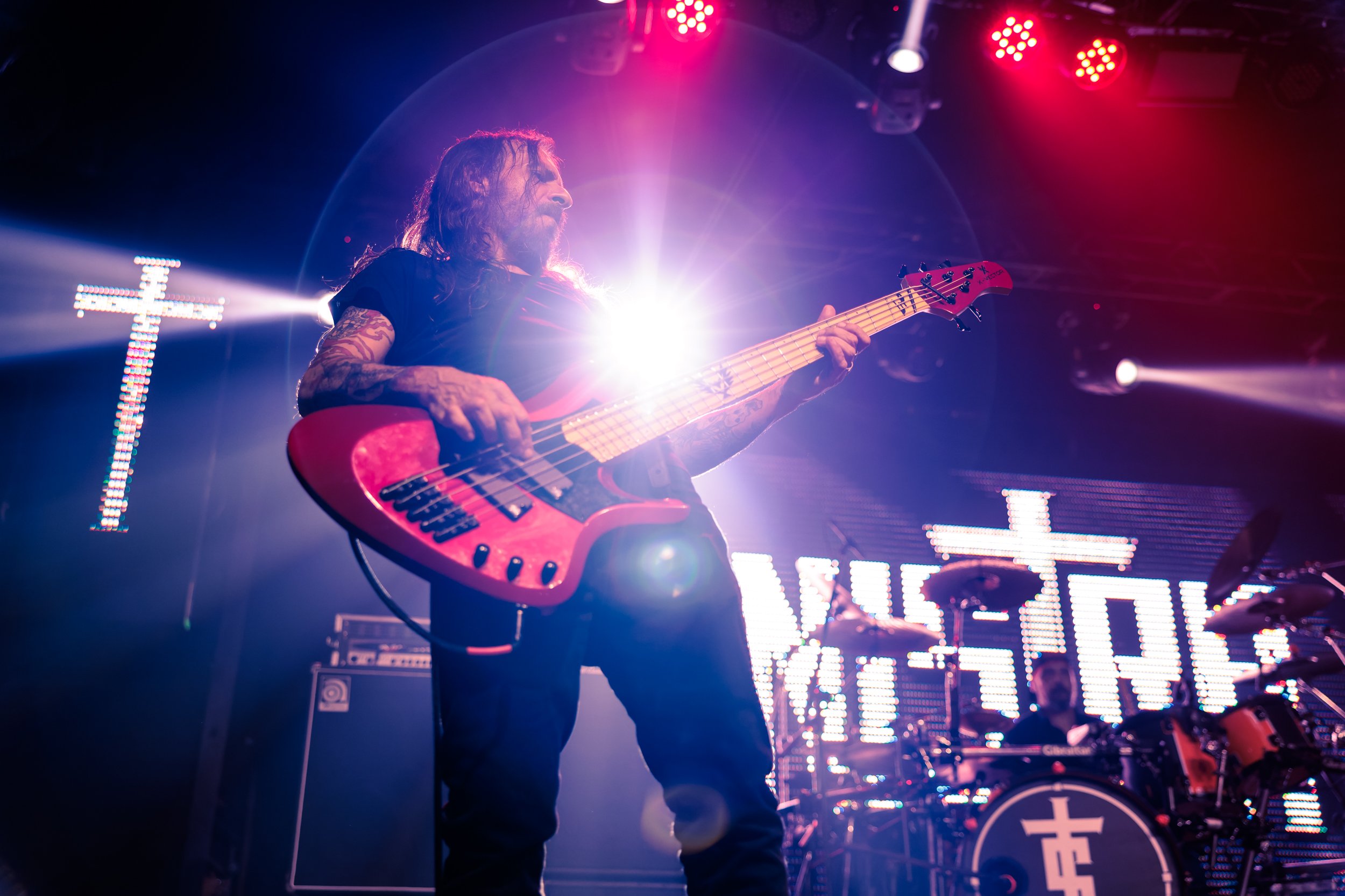 A musician with long hair and tattoos playing a red electric bass guitar on stage under colorful lights during a concert.