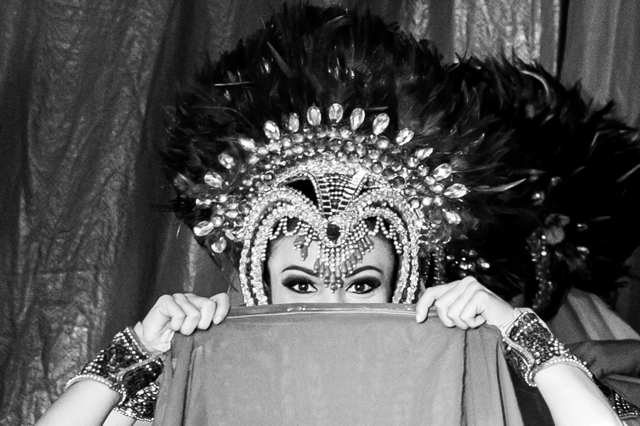 A woman with intricate beads and feathers on her head, holding a mirror in front of her face, showing her eyes, with her hands on each side of the mirror.