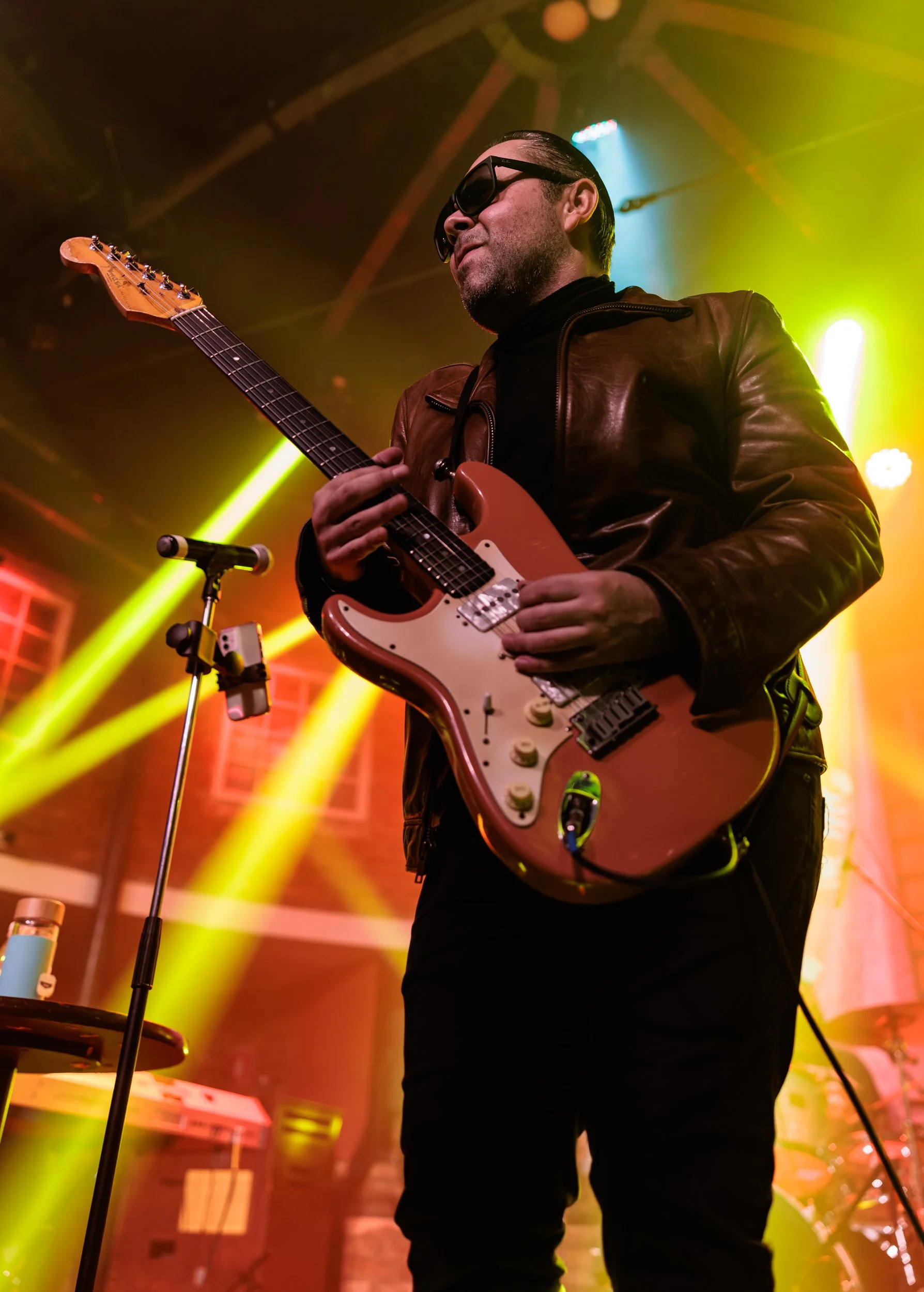 A man playing guitar on stage during a concert, wearing sunglasses and a brown leather jacket with colorful stage lighting.