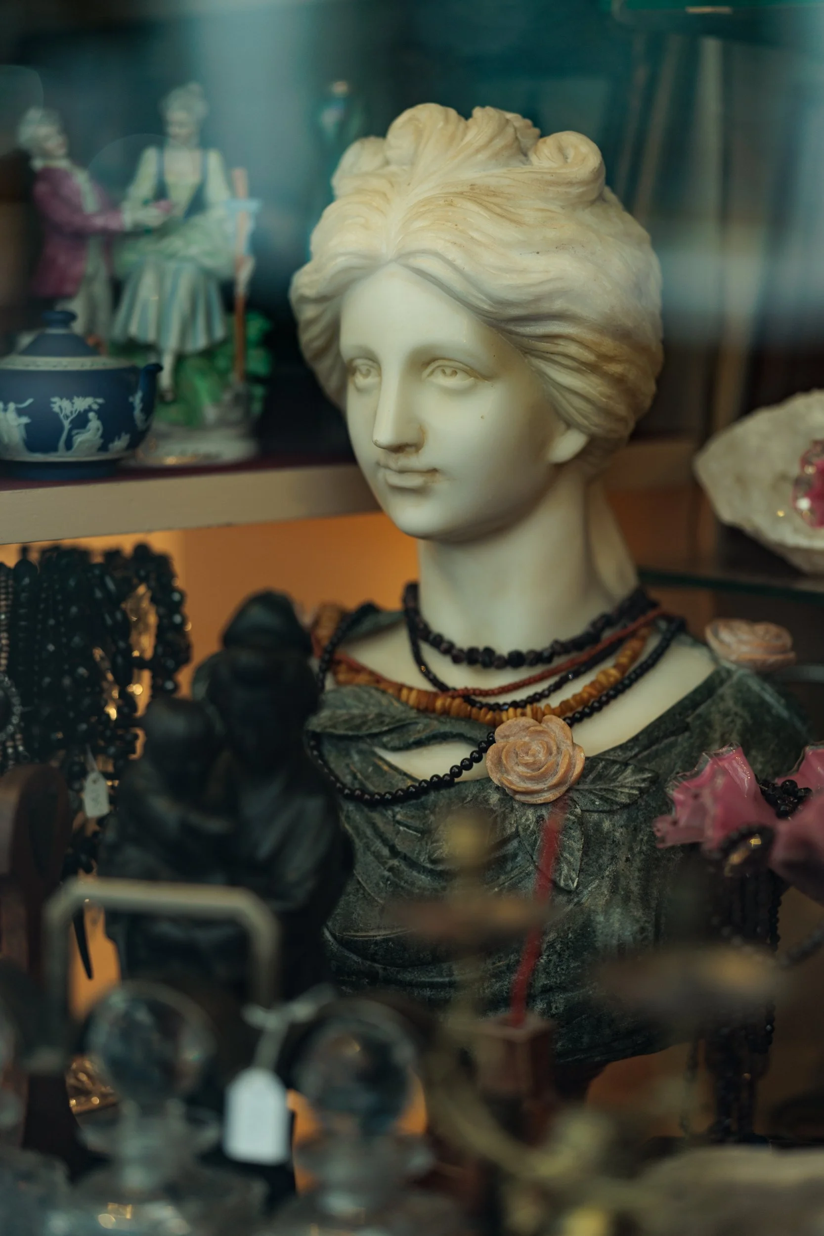 A porcelain bust of a woman with styled hair wearing multiple beaded necklaces, displayed behind glass alongside ceramic figurines and jewelry.