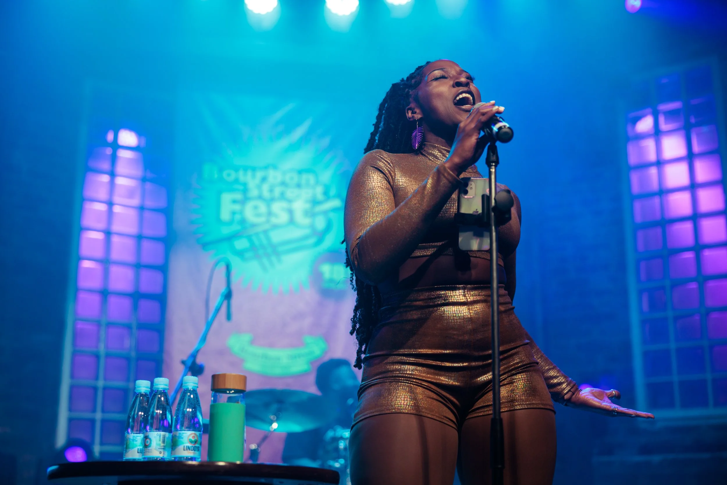 A woman singing passionately into a microphone on stage with colorful stage lights and a backdrop with a logo. She is wearing a shiny, textured outfit and large earrings.