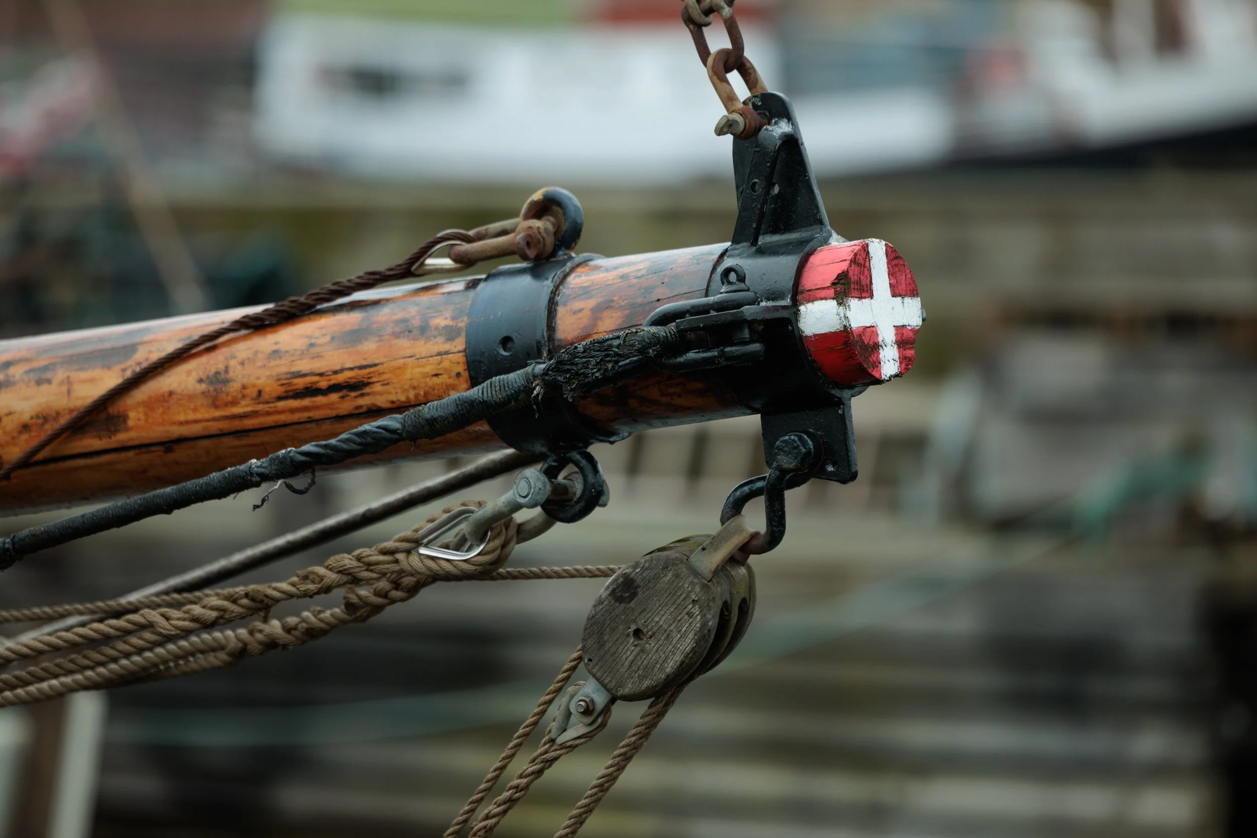Close-up of a boat's bow with a wooden pine, black metal fittings, and a painted red and white cross on the tip, with various ropes and pulleys attached.