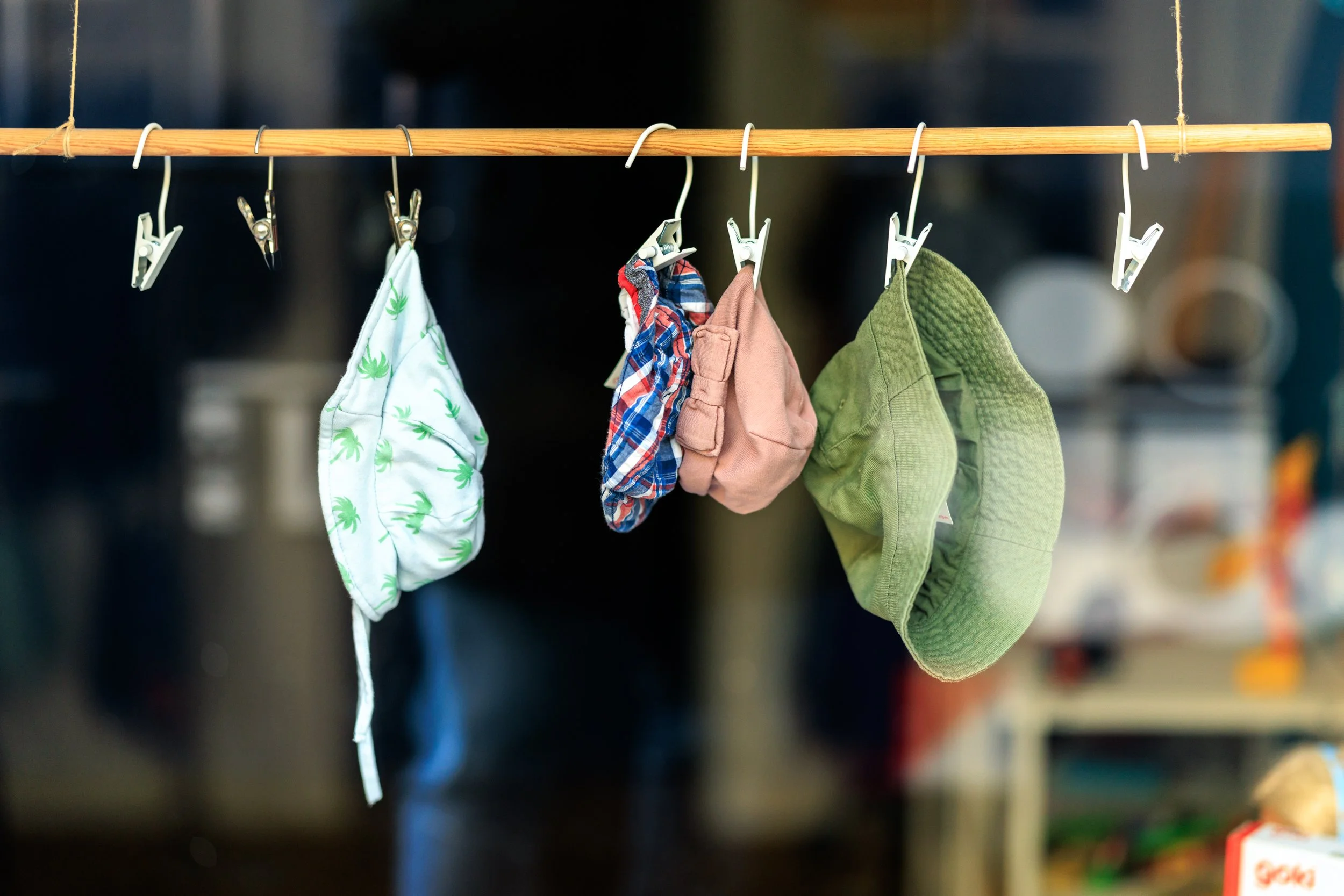 Clothes and fabric items hanging on a wooden rod with white clips, including a white towel with green leaf patterns, a plaid shirt, a pink cloth, and green quilted fabric, in a retail or storage setting.