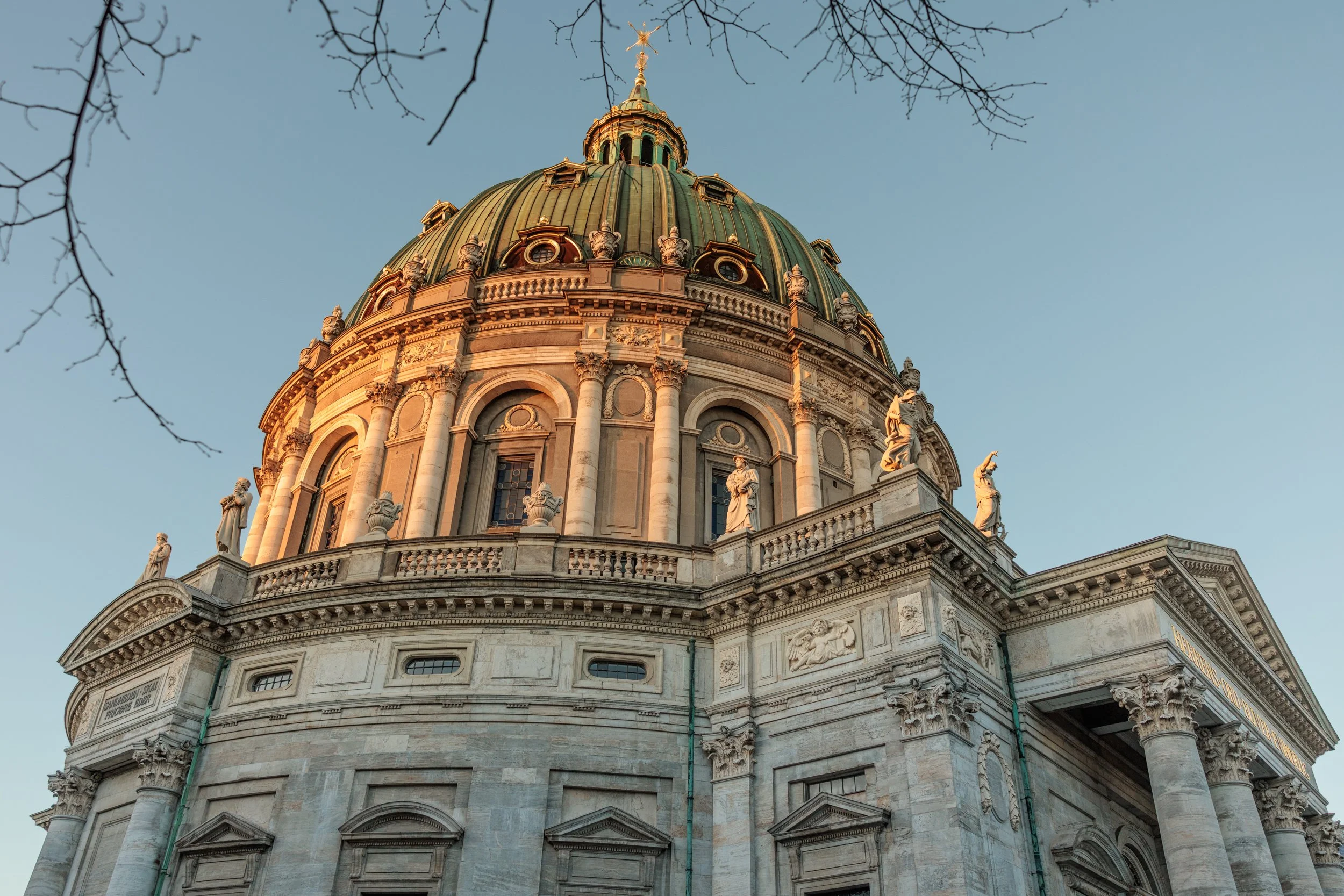 The dome of a historic building with statues along the roofline, seen against a clear sky, with leafless tree branches at the top.