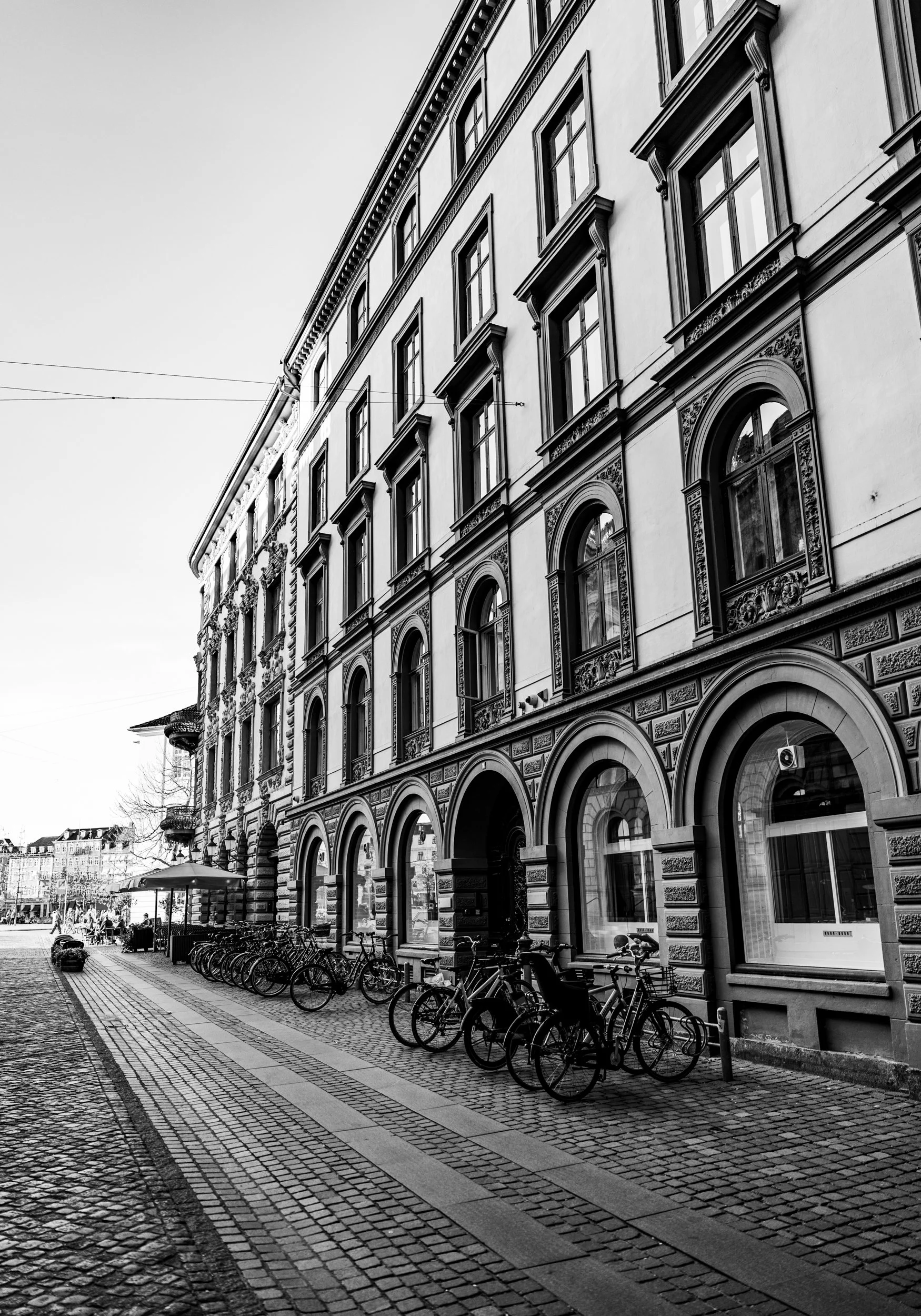 Black and white photo of a historic building with decorative architectural details, arched windows, and multiple stories. Bicycles are parked along the sidewalk, and outdoor seating with umbrellas is visible. Overhead wires cross the street, indicati