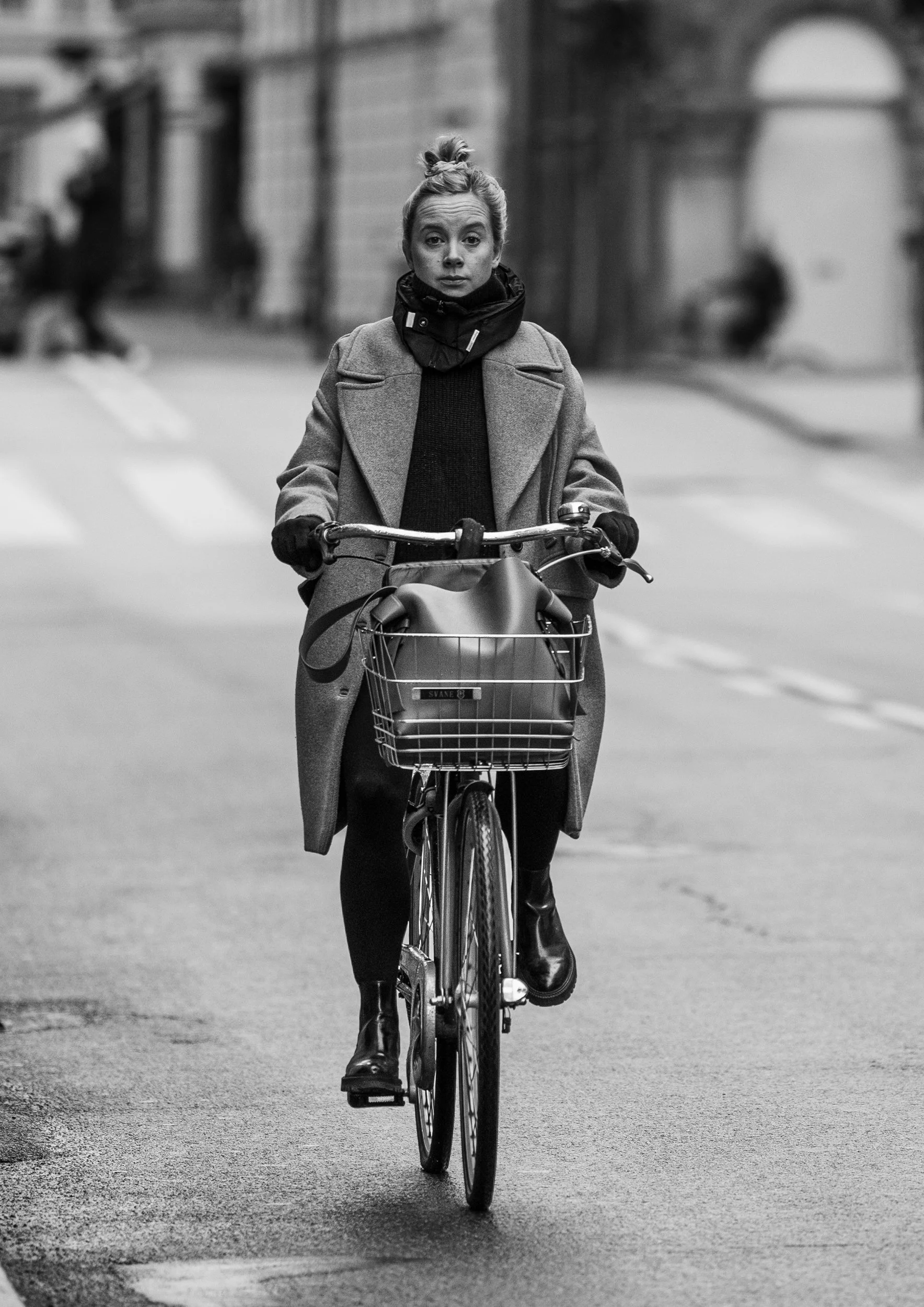 A woman with blonde hair tied in a bun, wearing a coat, gloves, and a scarf, is riding a bicycle on a city street. The background shows blurred buildings and pedestrians.