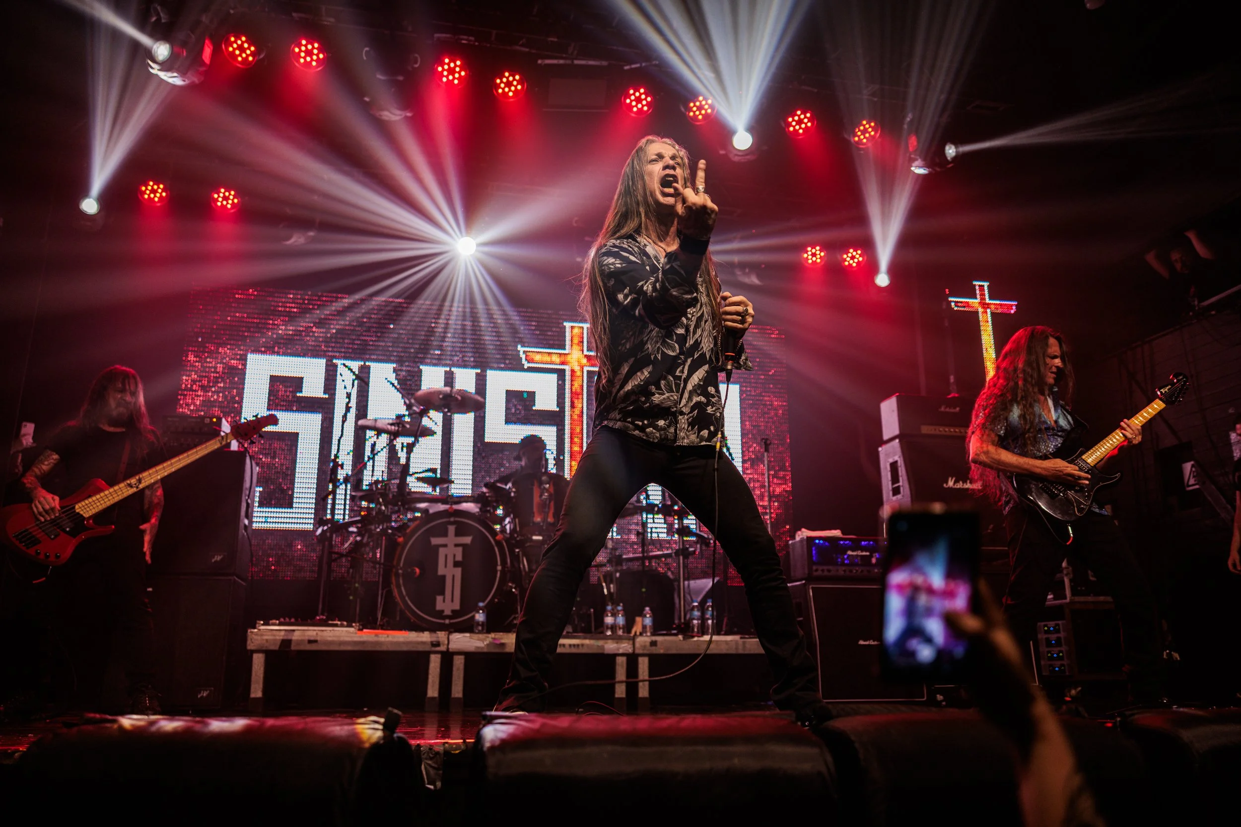 A band performing on stage with a female lead singer making an expressive gesture and singing, illuminated by bright red and white stage lights with a LED screen displaying text and crosses in the background.