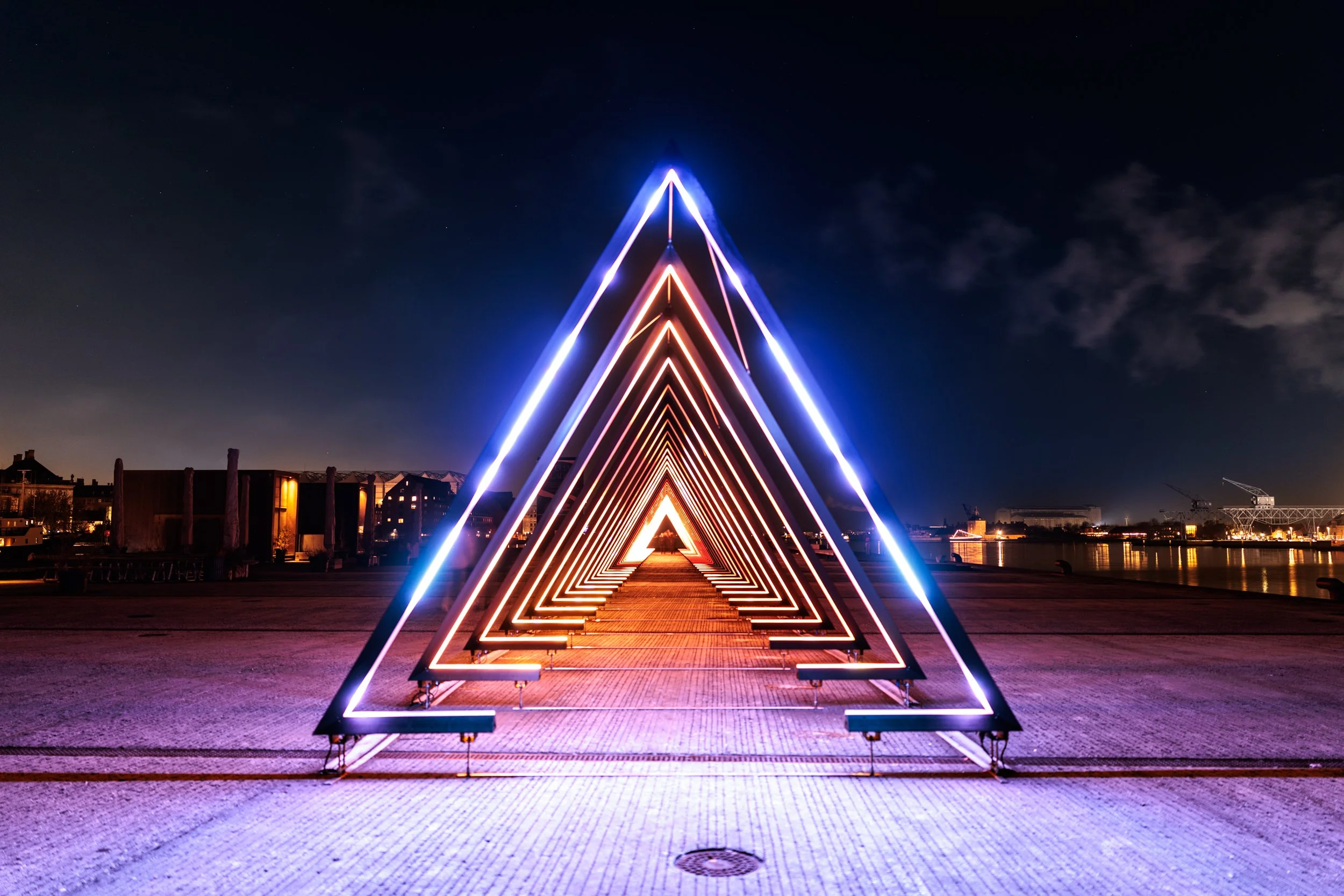 Night view of illuminated triangular-shaped neon light installation on a deck with a cityscape and river in the background.