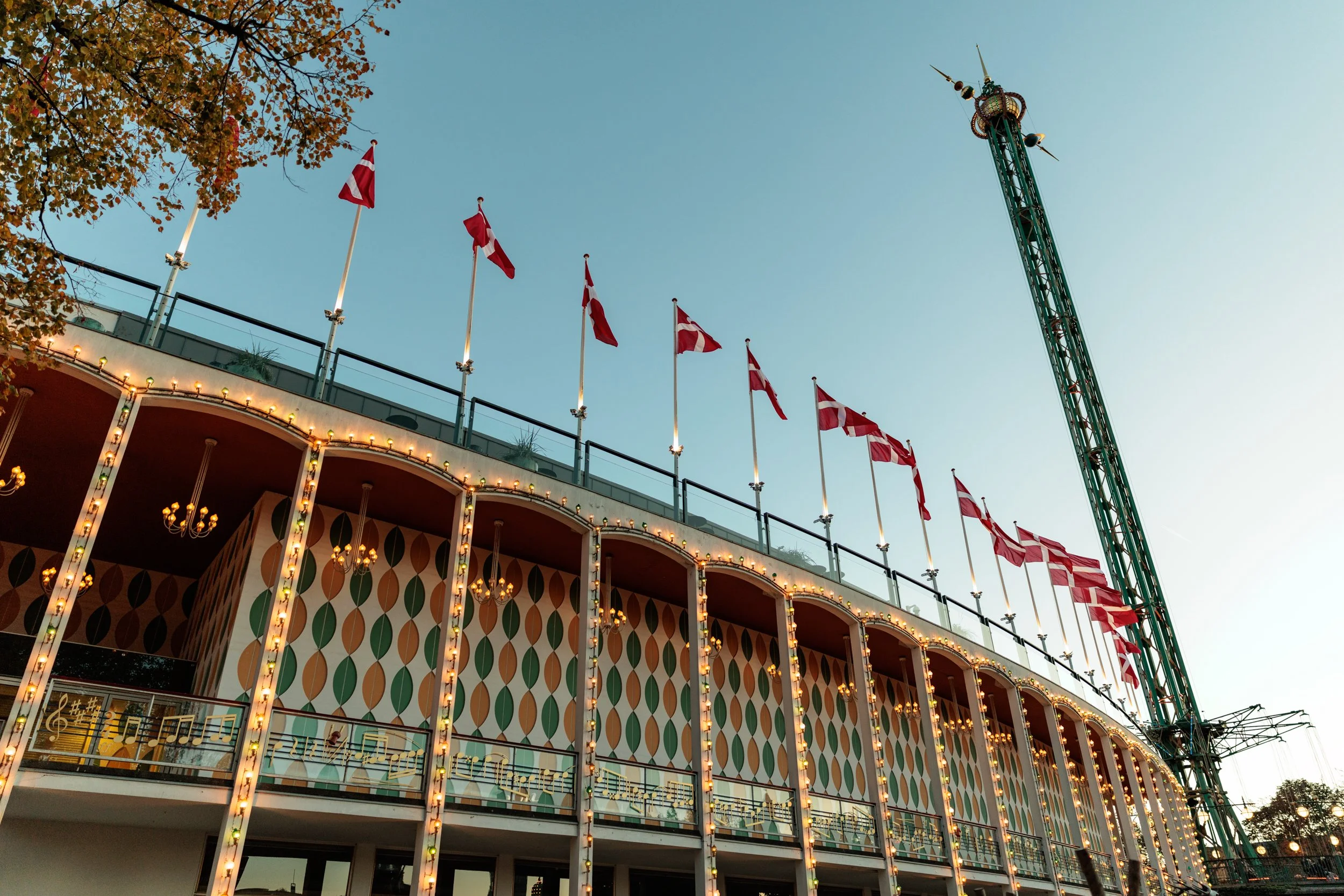 The image shows a section of a fairground with a building decorated with colorful diamond-shaped patterns and string lights hanging from its edges. Several Danish flags are visible on the building's balcony, and a tall amusement ride tower is seen ex