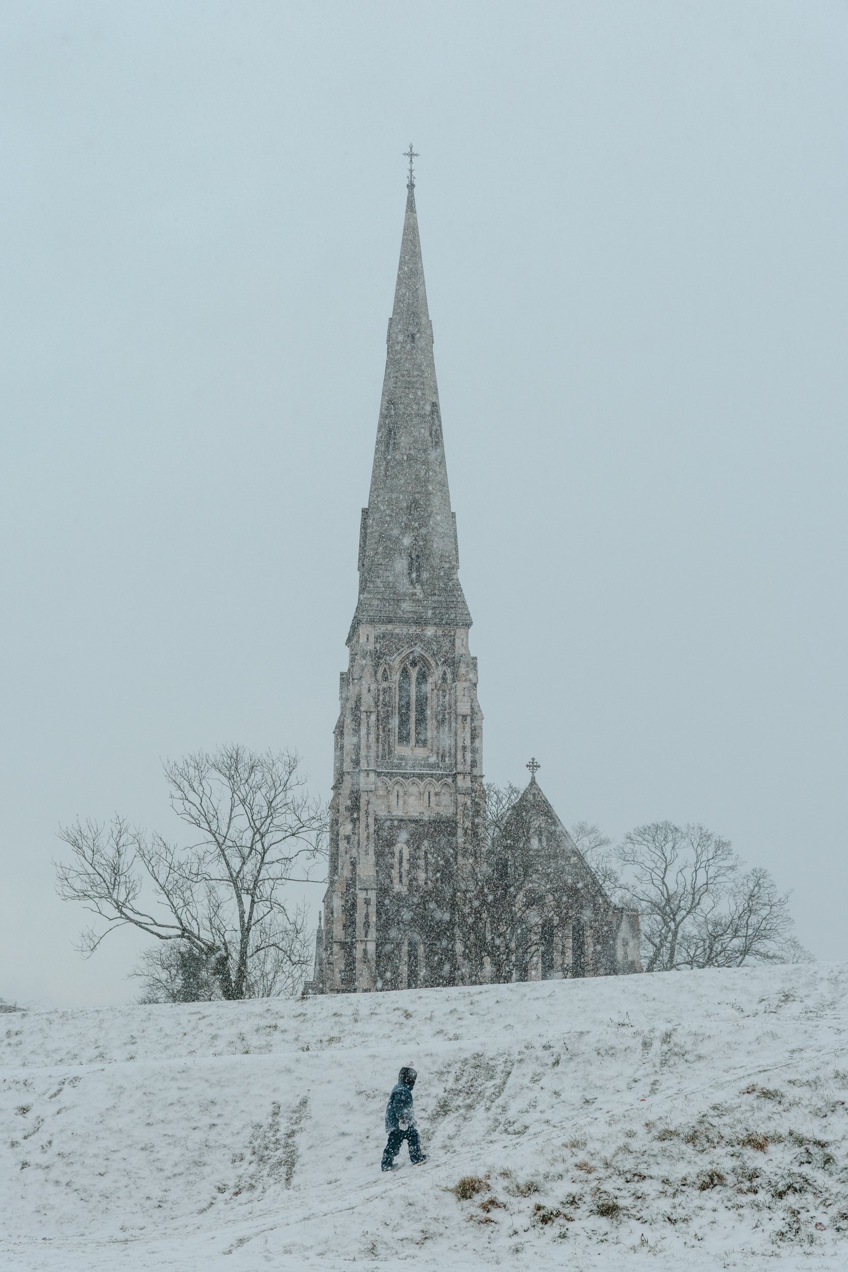 A snowy scene featuring a large church with a tall steeple topped with a cross. Snow is falling heavily, and a person in dark clothing is walking up a snow-covered hill in front of the church, with leafless trees surrounding the church.