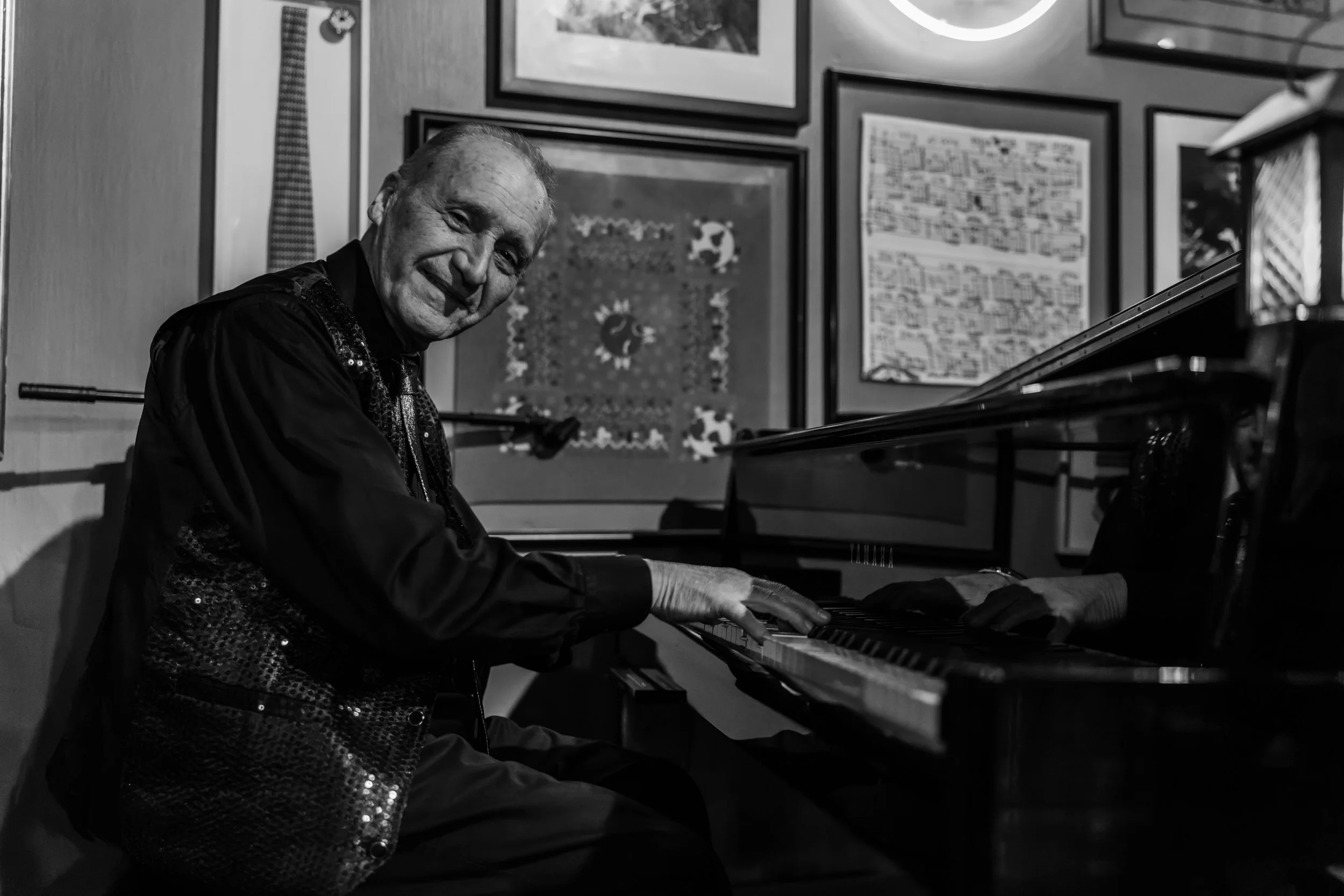 An elderly man smiling while playing the piano in a room decorated with framed artwork on the wall.