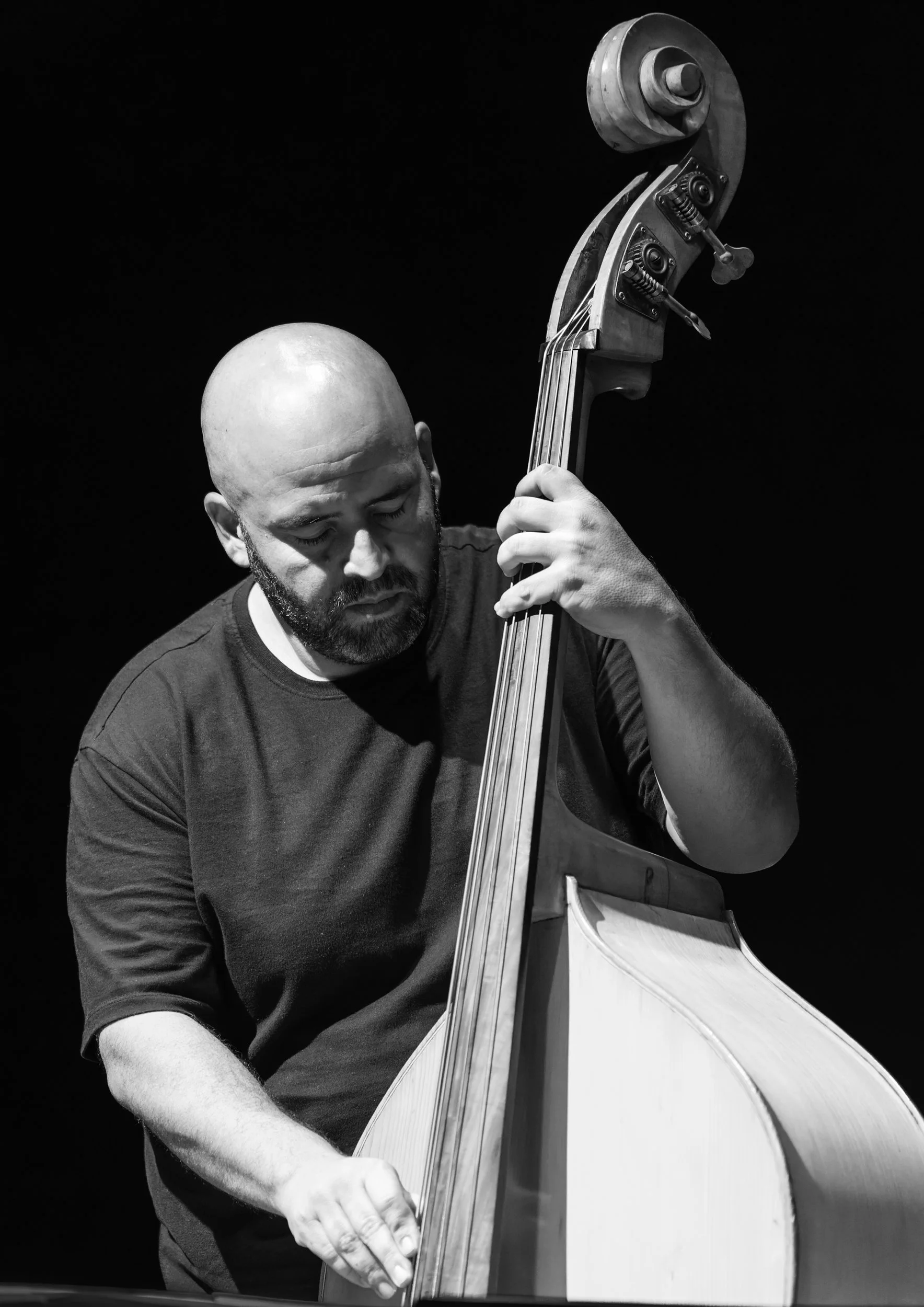 Black and white photo of a man with a shaved head playing a double bass, with focus on his face and instrument against dark background.
