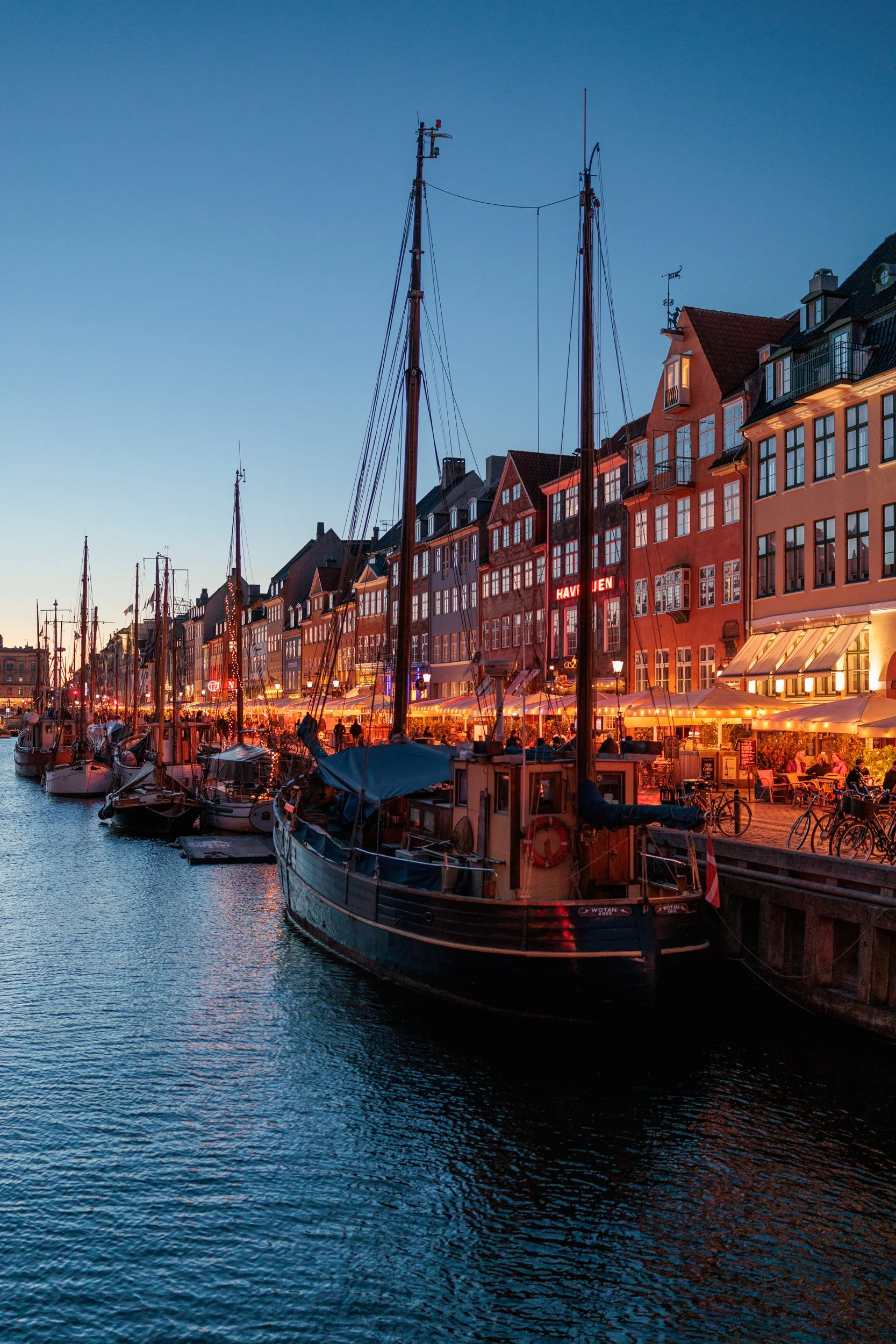 Boats docked along a waterfront at dusk with illuminated buildings and outdoor seating area, capturing a lively harbor scene.