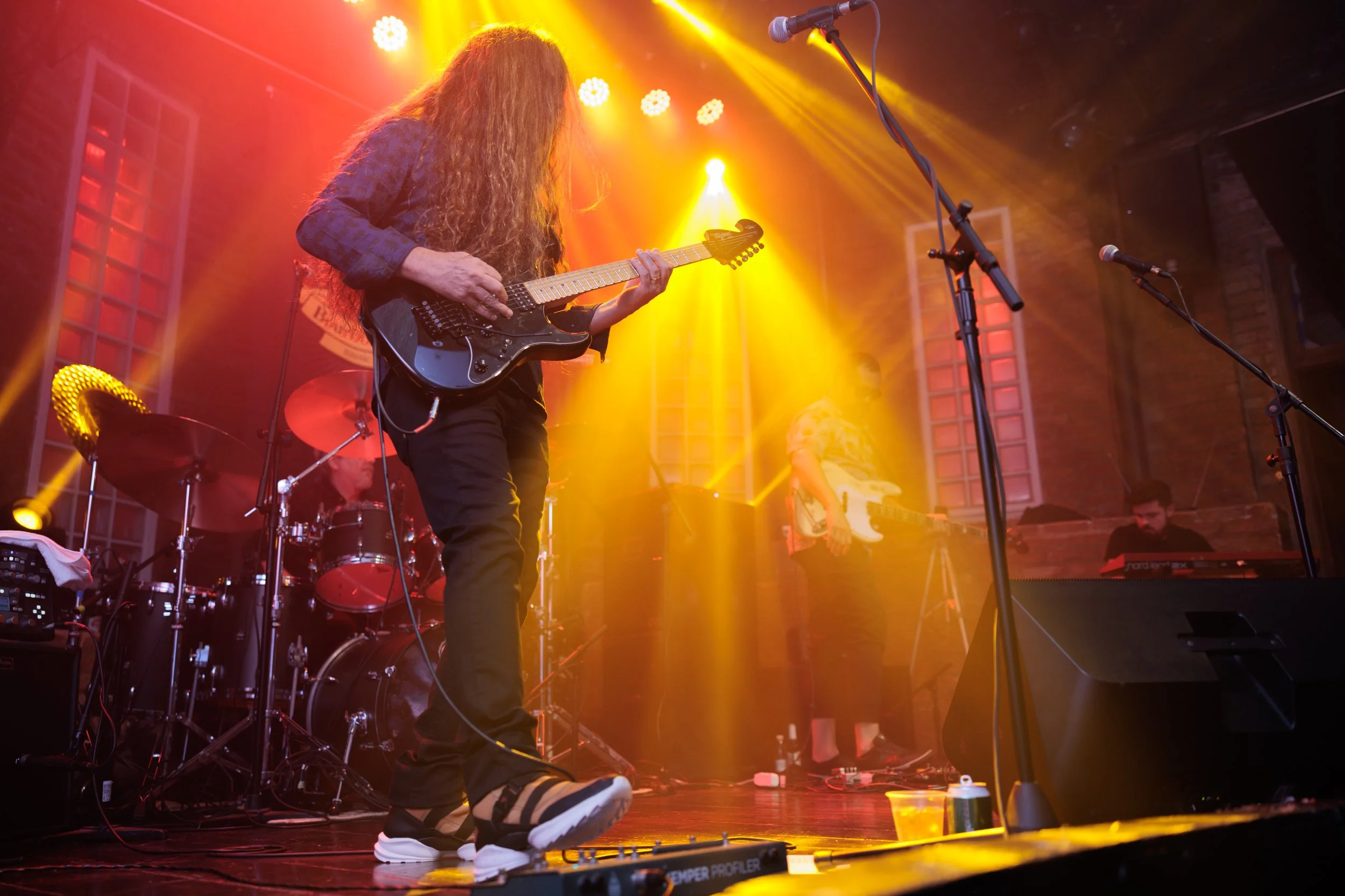 Musician with long curly hair playing an electric guitar on stage illuminated by yellow and orange stage lights, with band members and musical equipment visible in the background.