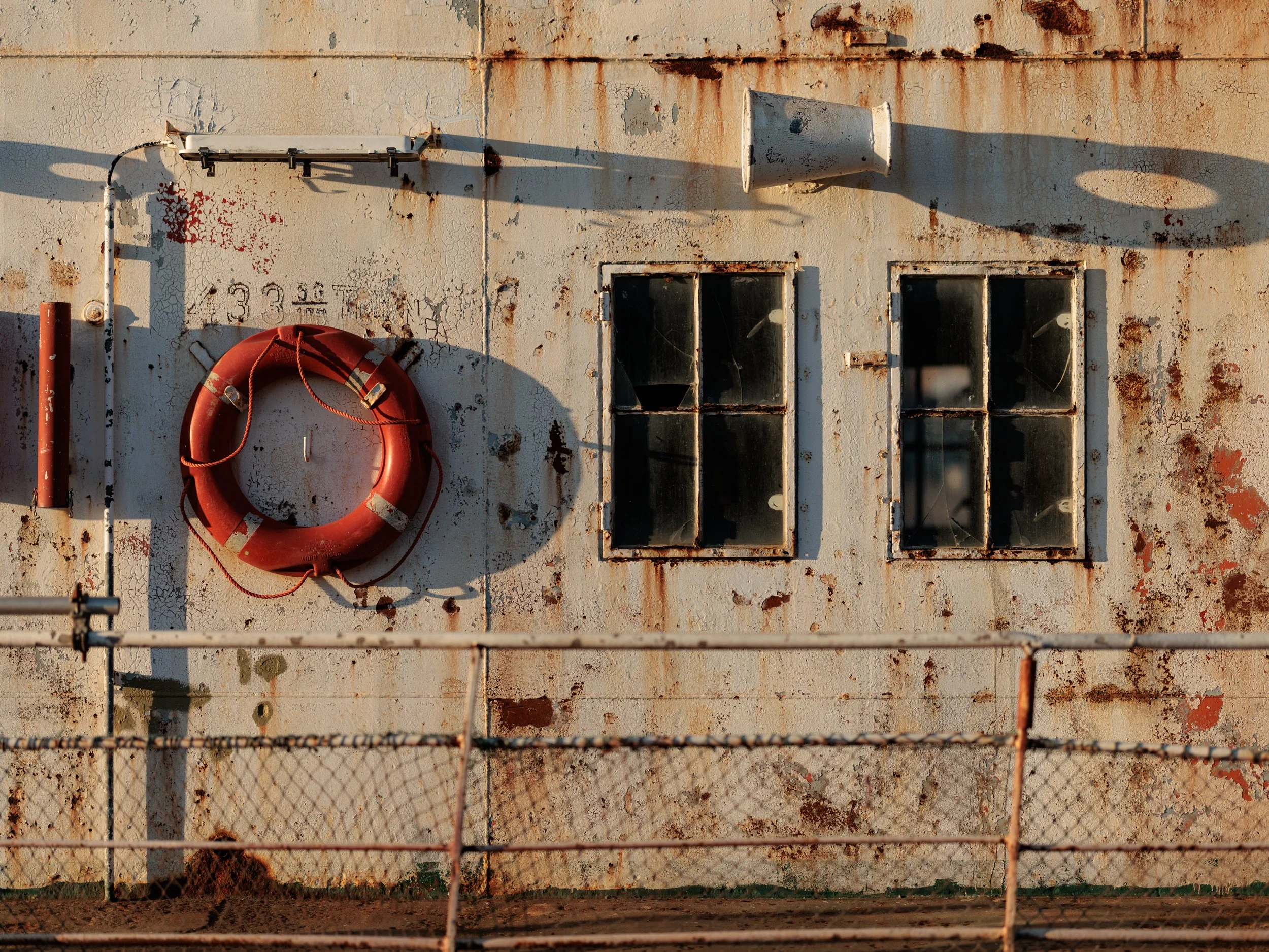 A weathered, rusted metal wall with two small, dirty windows, a life preserver hanging on a yellow pipe, and an outdoor loudspeaker. A chain-link fence partially obscures the lower part of the scene.