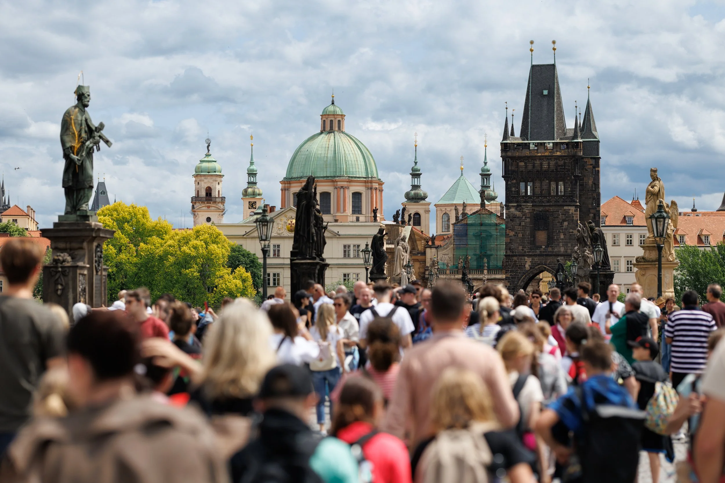 Crowd of people in a historic city square with statues, church domes, and clock tower in the background.