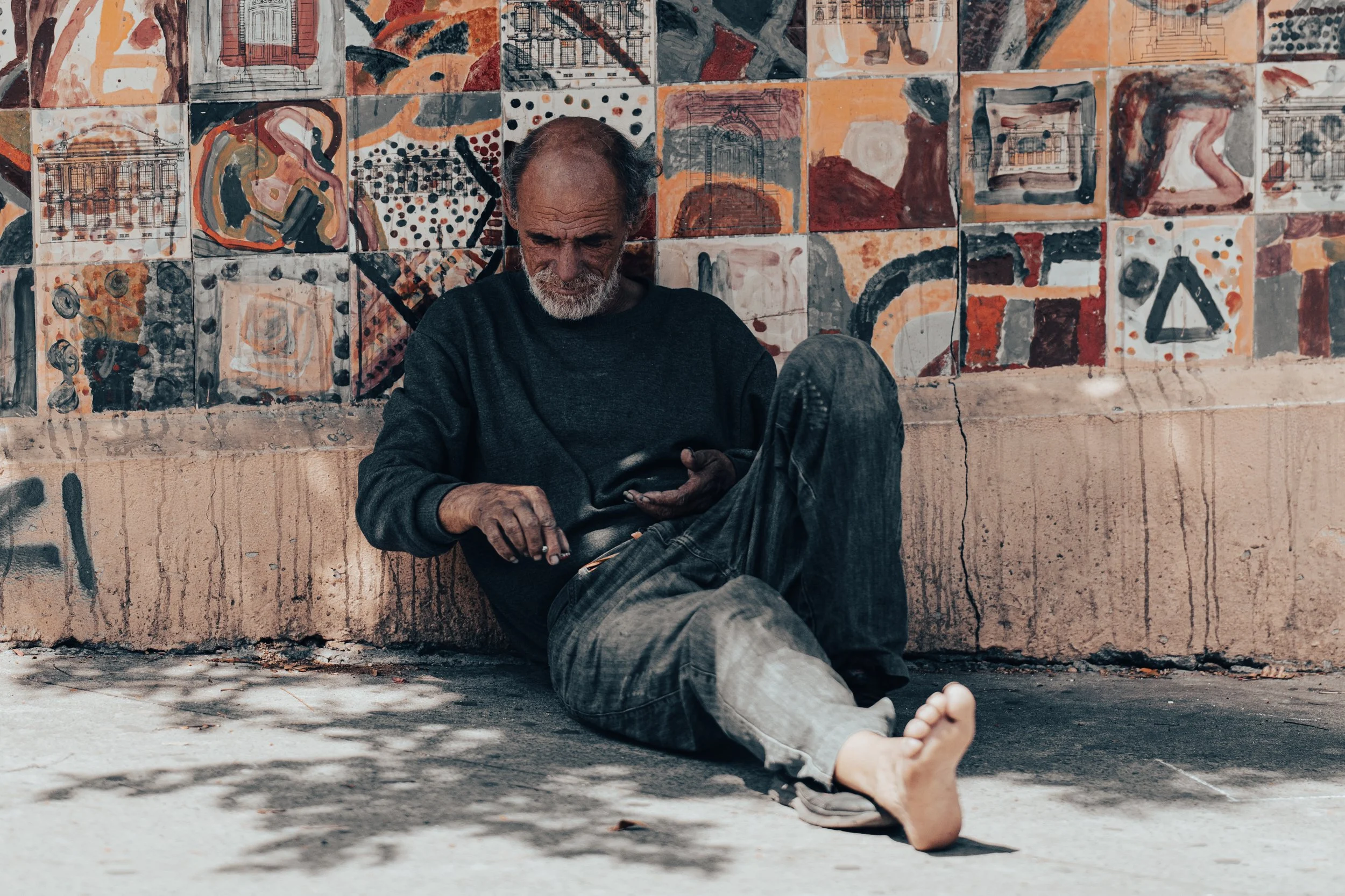 An elderly man with gray hair and beard sitting against a colorful mural, looking down at his phone. His attire is casual, wearing a dark long-sleeved shirt and jeans. His feet are bare, and he appears to be resting on a sidewalk.
