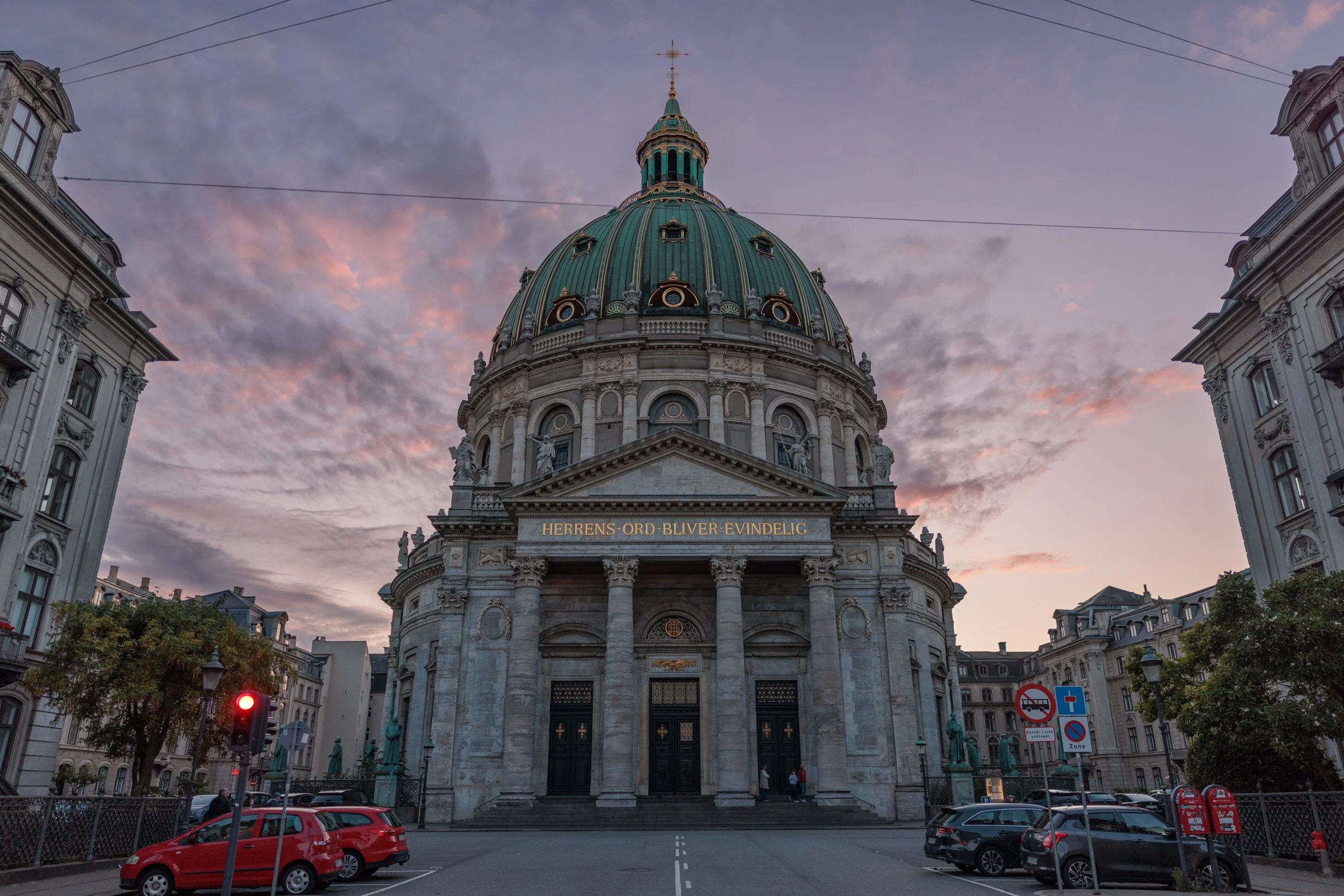 A historic building with a large green dome, columns, and statues at the entrance, under a sky with pink and purple clouds, surrounded by cars and street signs.