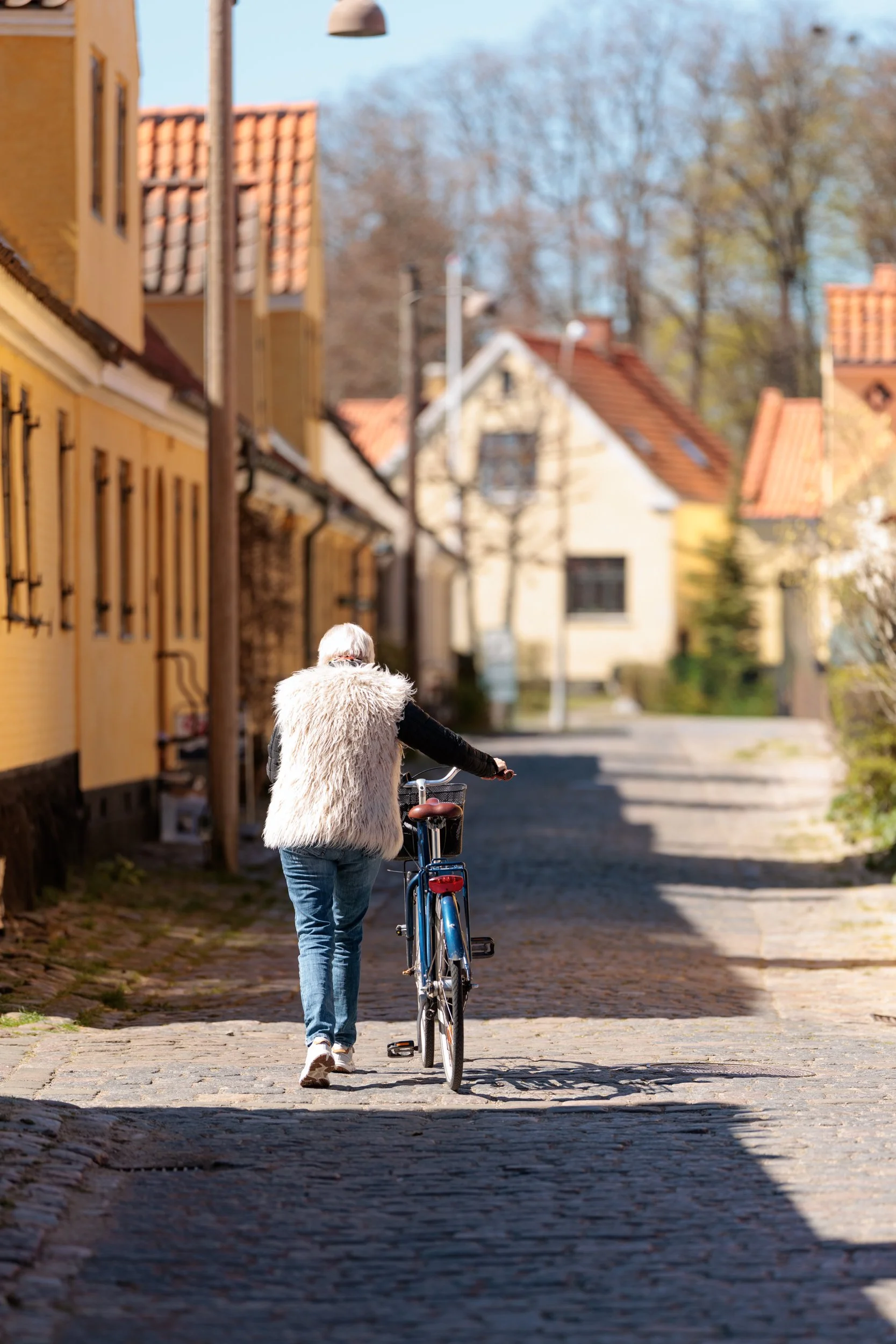 A person with white hair, wearing a fluffy vest, walking a bicycle down a cobblestone street lined with colorful houses on a sunny day.