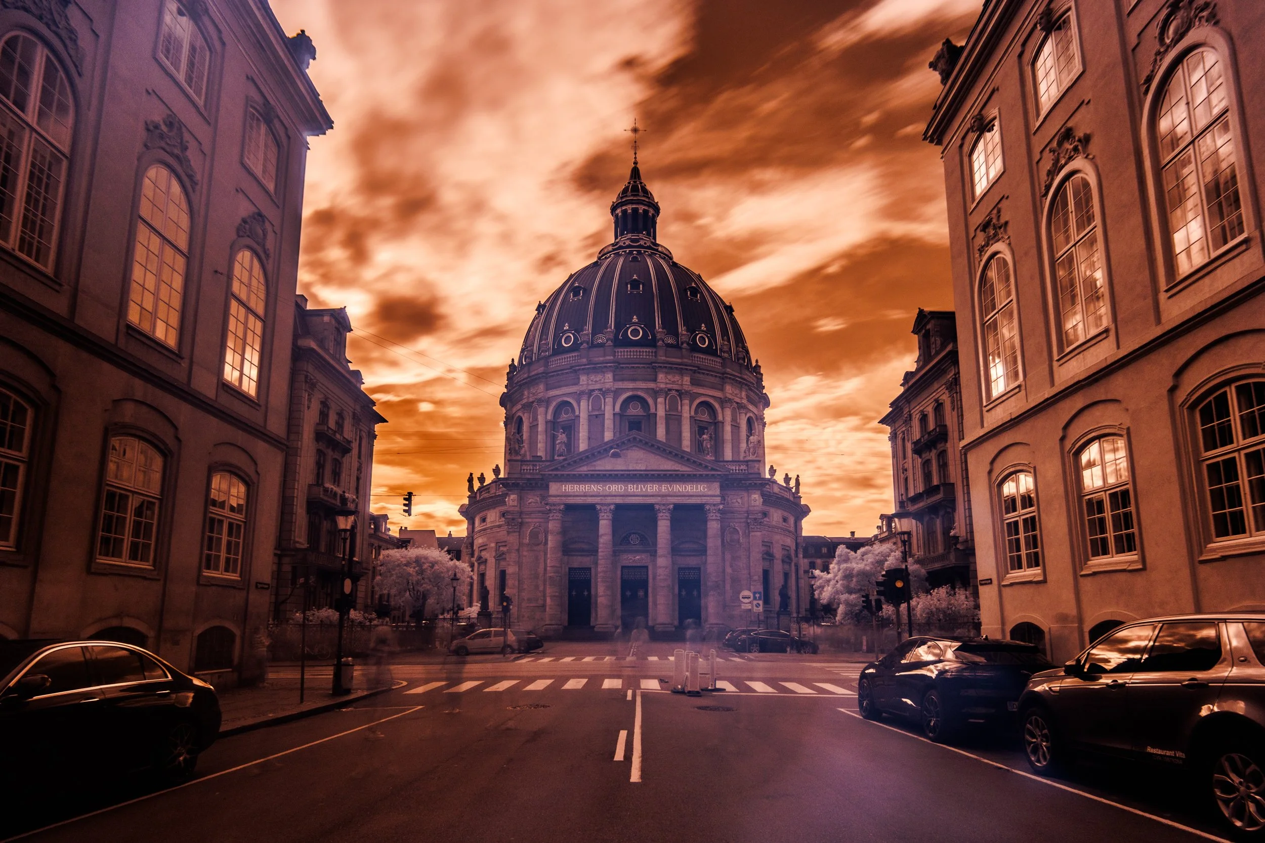 Photo of a historic building with a large dome, surrounded by classical architecture, under an orange cloudy sky at sunset.