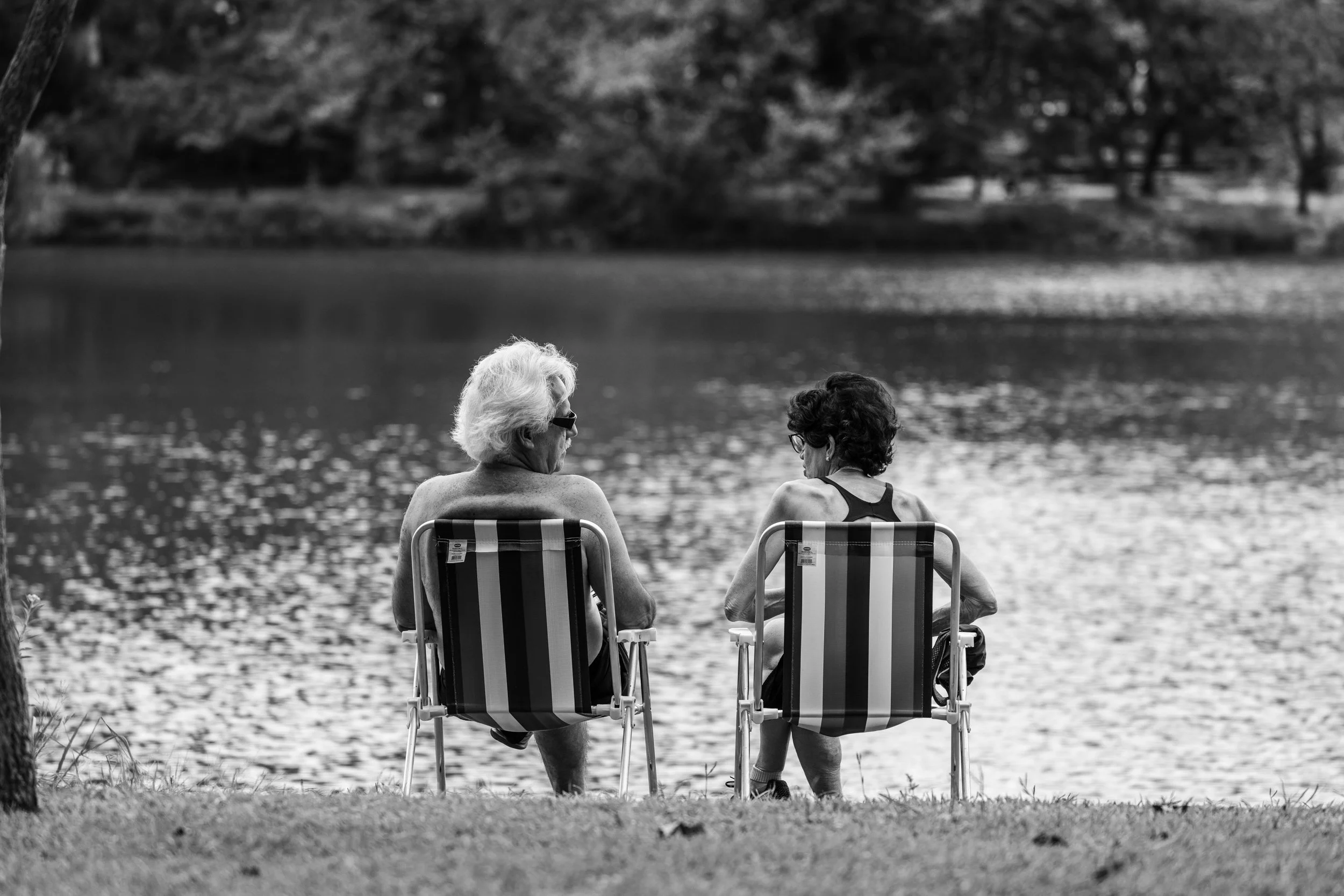 Two elderly women sitting on striped lawn chairs by a lake, facing away from the camera, engaging in conversation, with trees across the water in the background.