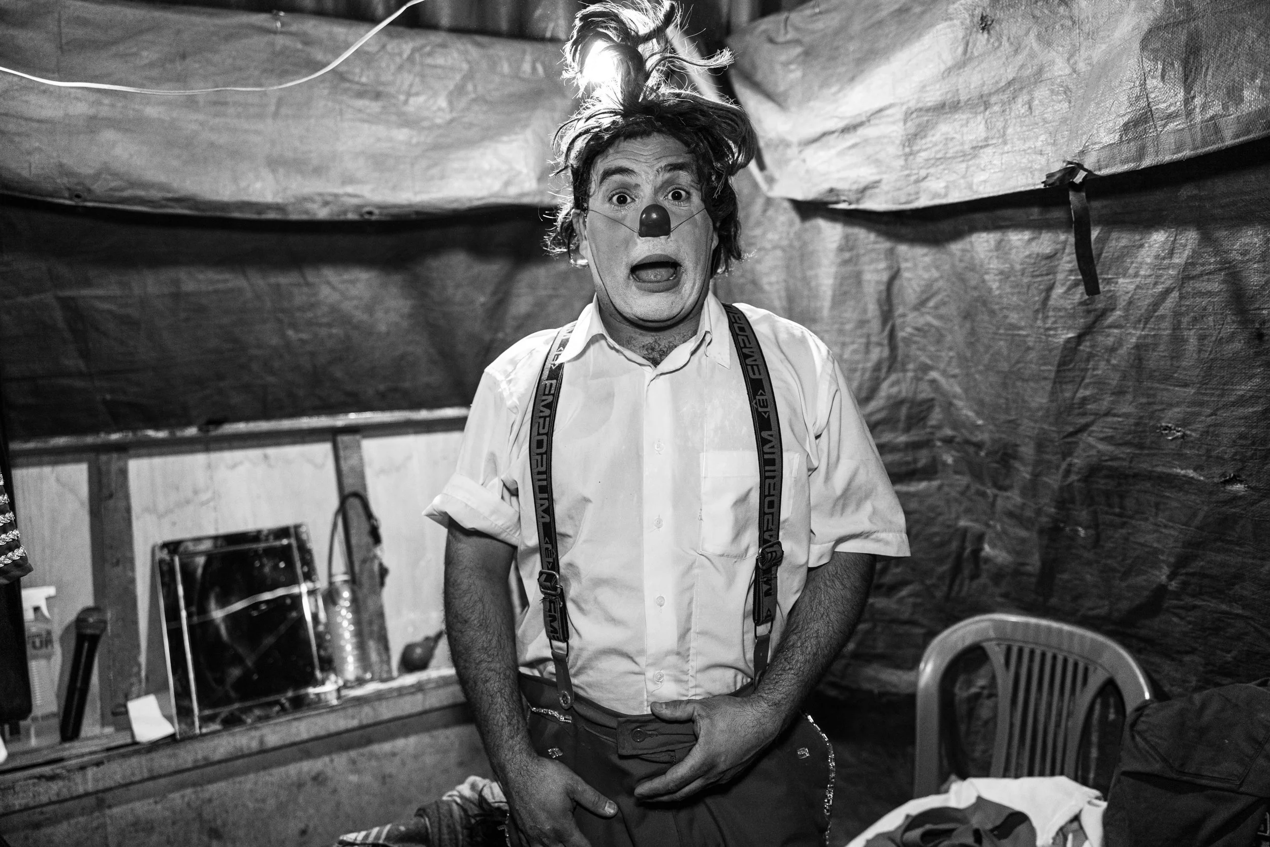 A person dressed as a clown with a red nose, wild hair, and suspenders, standing in an indoor setting with a surprised or shocked expression.