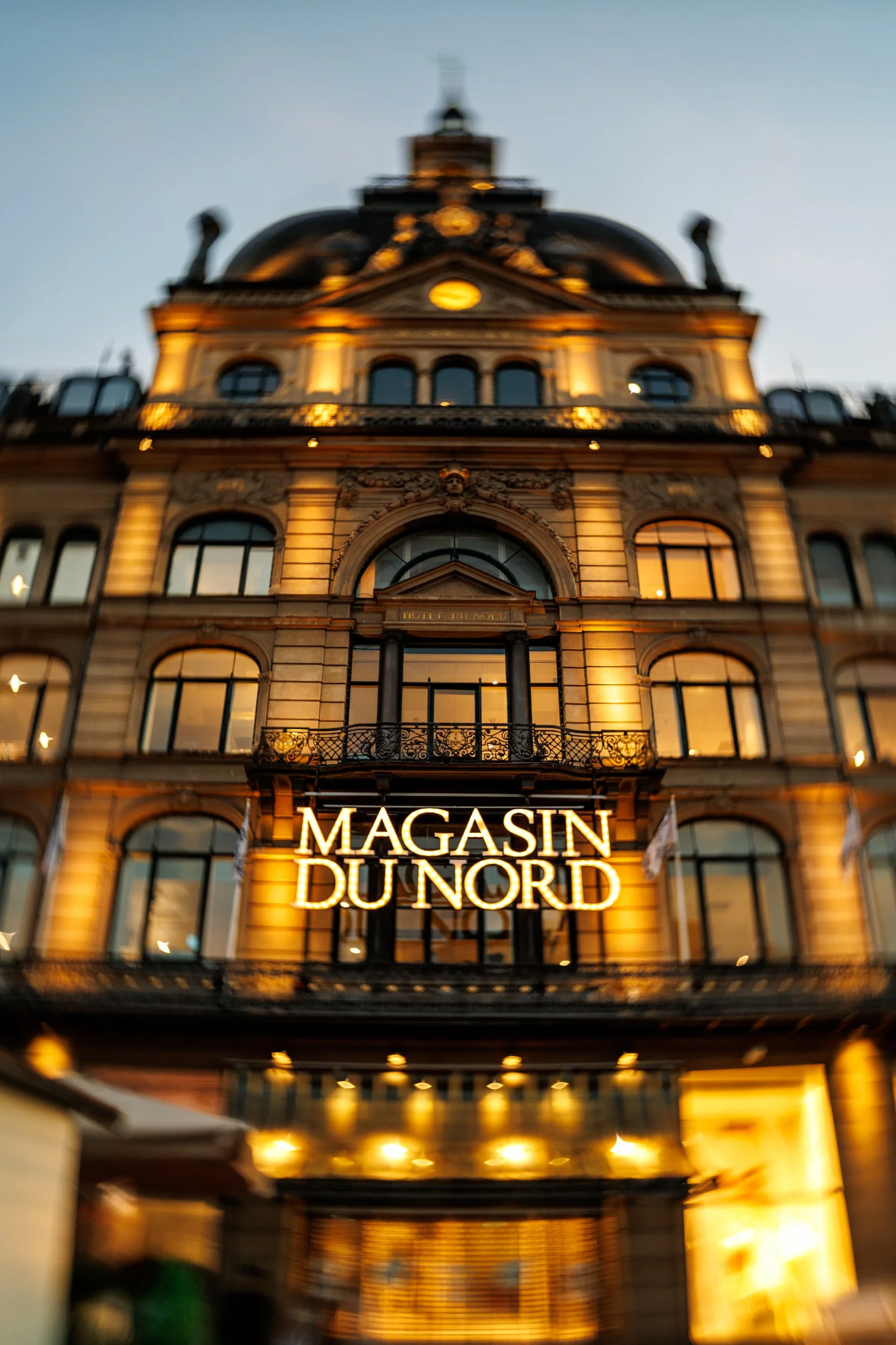 A historic multi-story building with ornate architectural details, illuminated at night. A bright neon sign at the entrance reads "MAGASIN DUNORD."