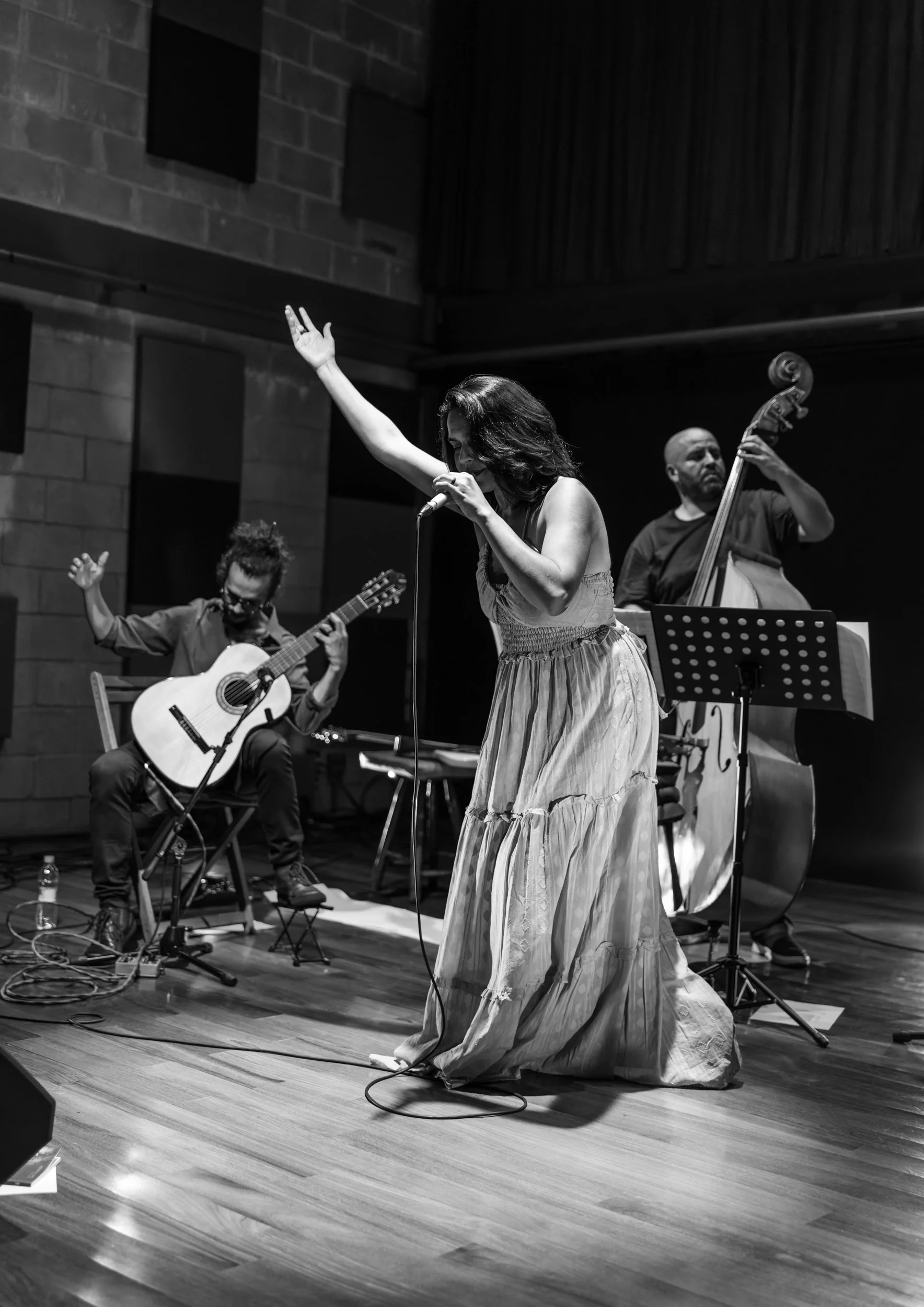 A black and white photo of a woman singing into a microphone on stage, with a man playing guitar and another man playing double bass in the background.