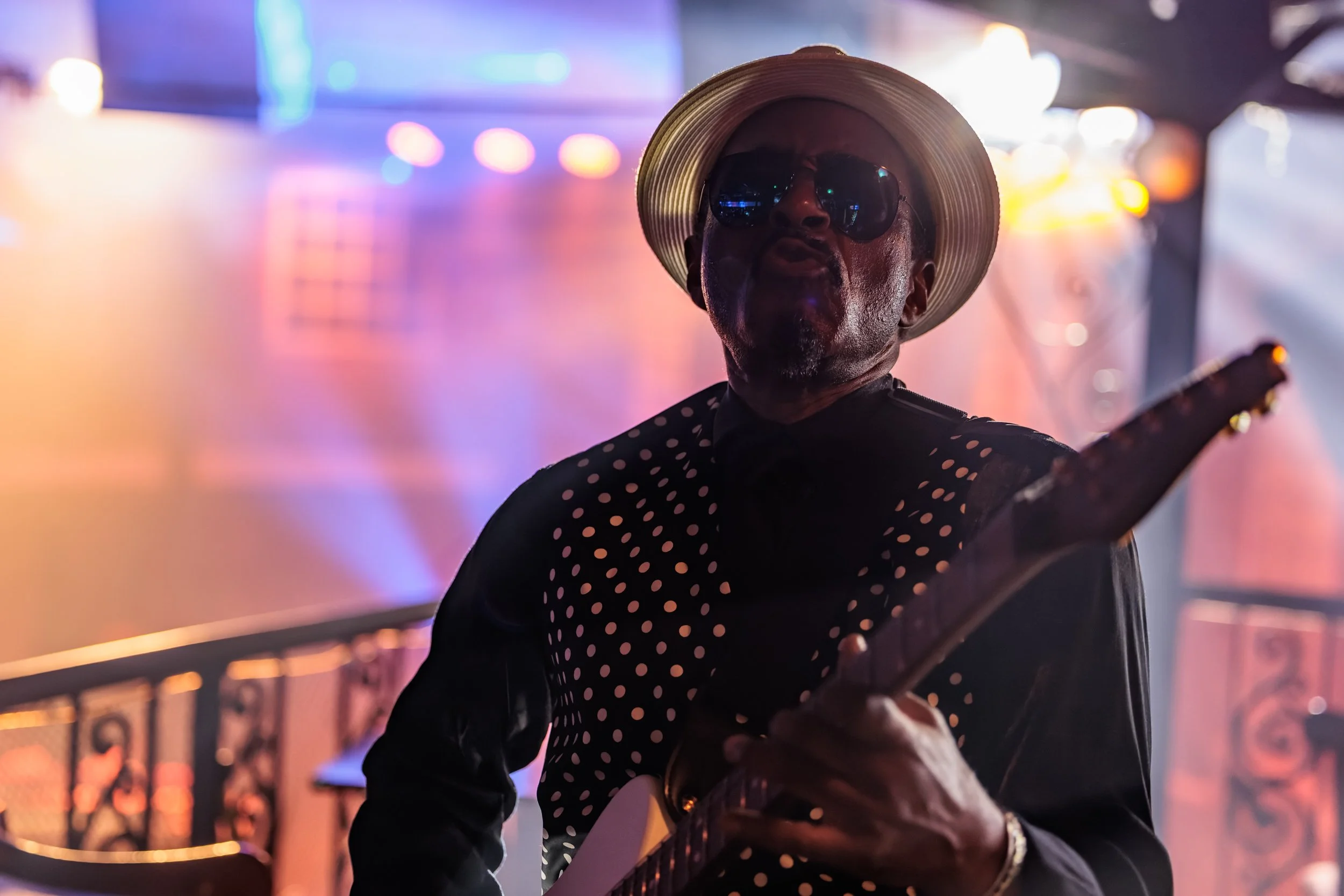 A man wearing a wide-brimmed hat, sunglasses, and a polka-dot shirt playing a guitar indoors with colorful blurred lights in the background.